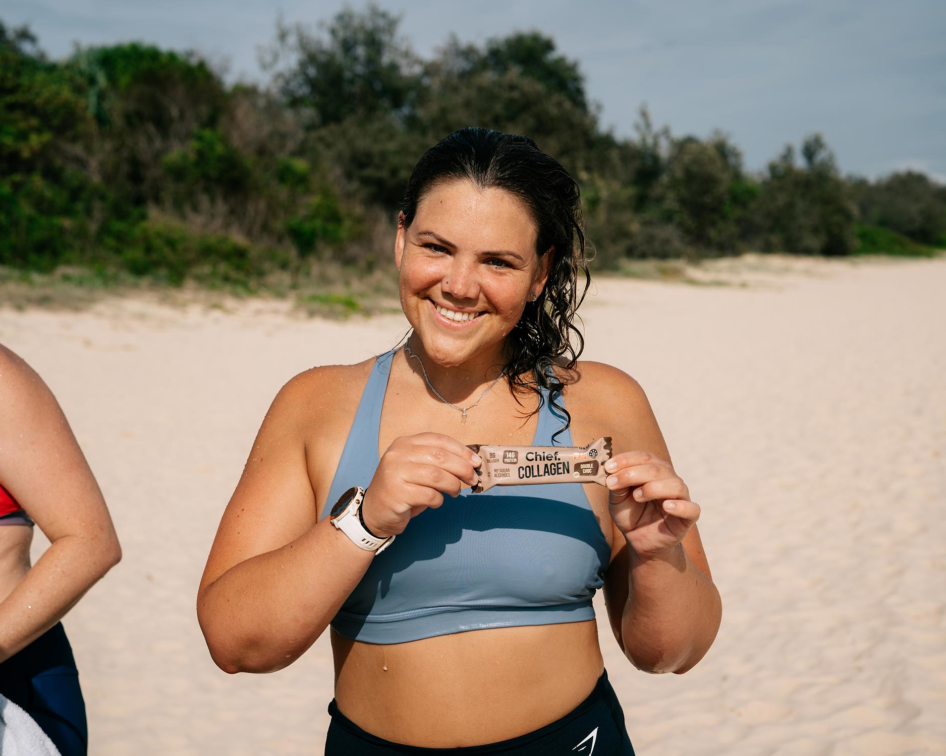 A woman on the beach in activewear smiling at the camera and holding up a Chief Collagen Bar