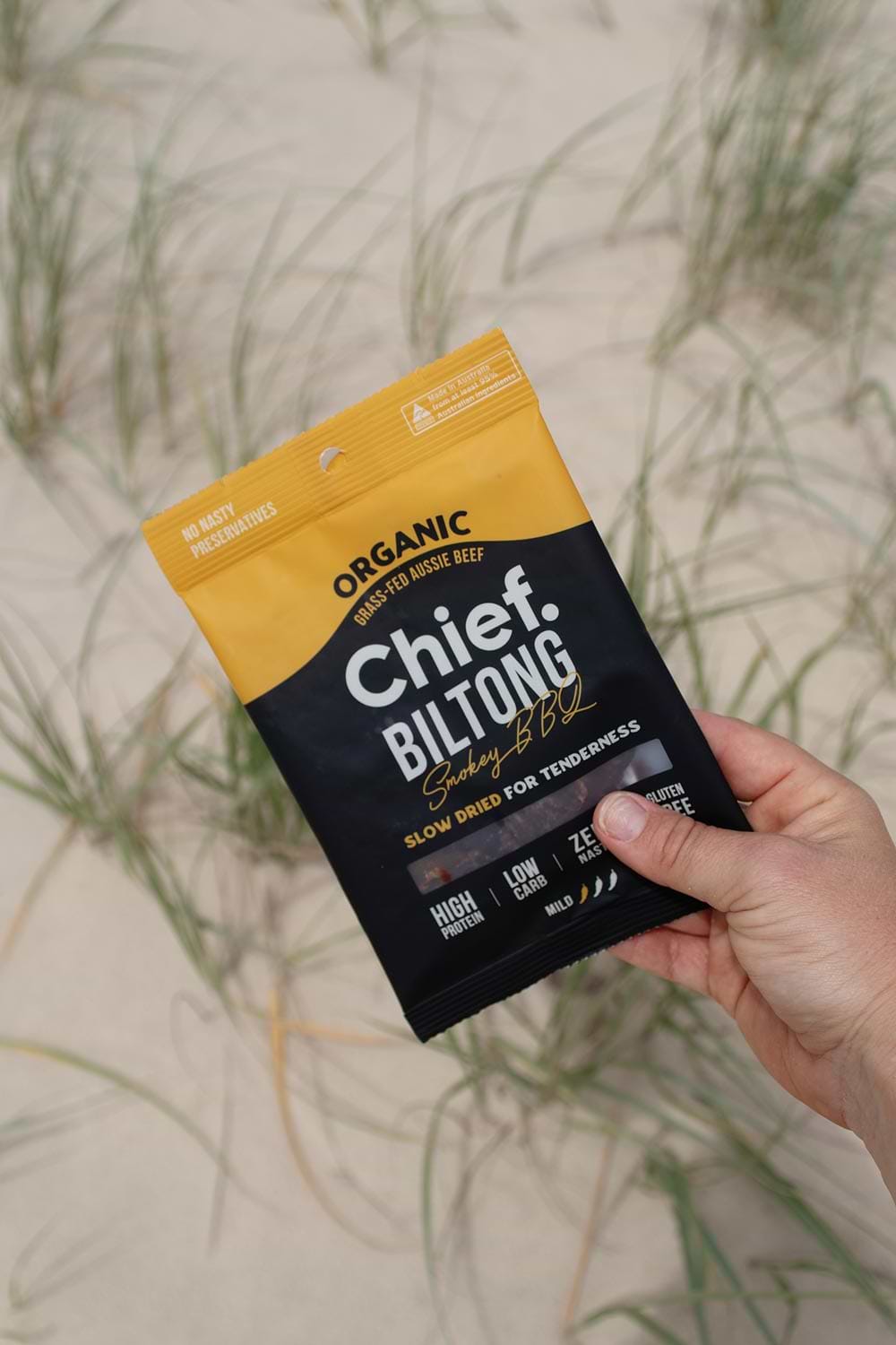 A person's hand holding a bag of Chief Smokey BBQ Biltong against a sand dune backdrop