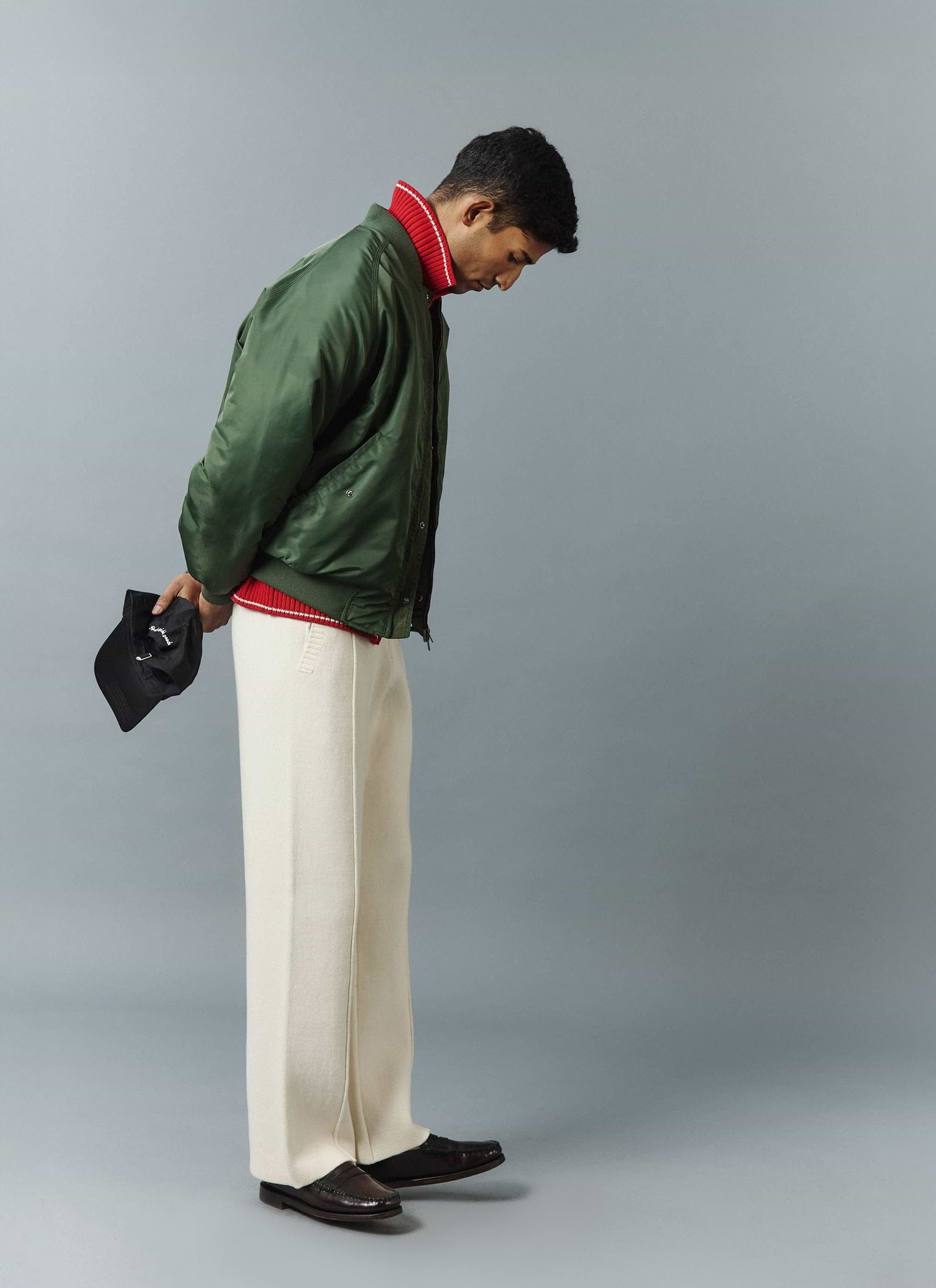 A model, shot from the side, wears the Percival Nylon Bomber Jacket in Forest over a red knit and cream trousers.