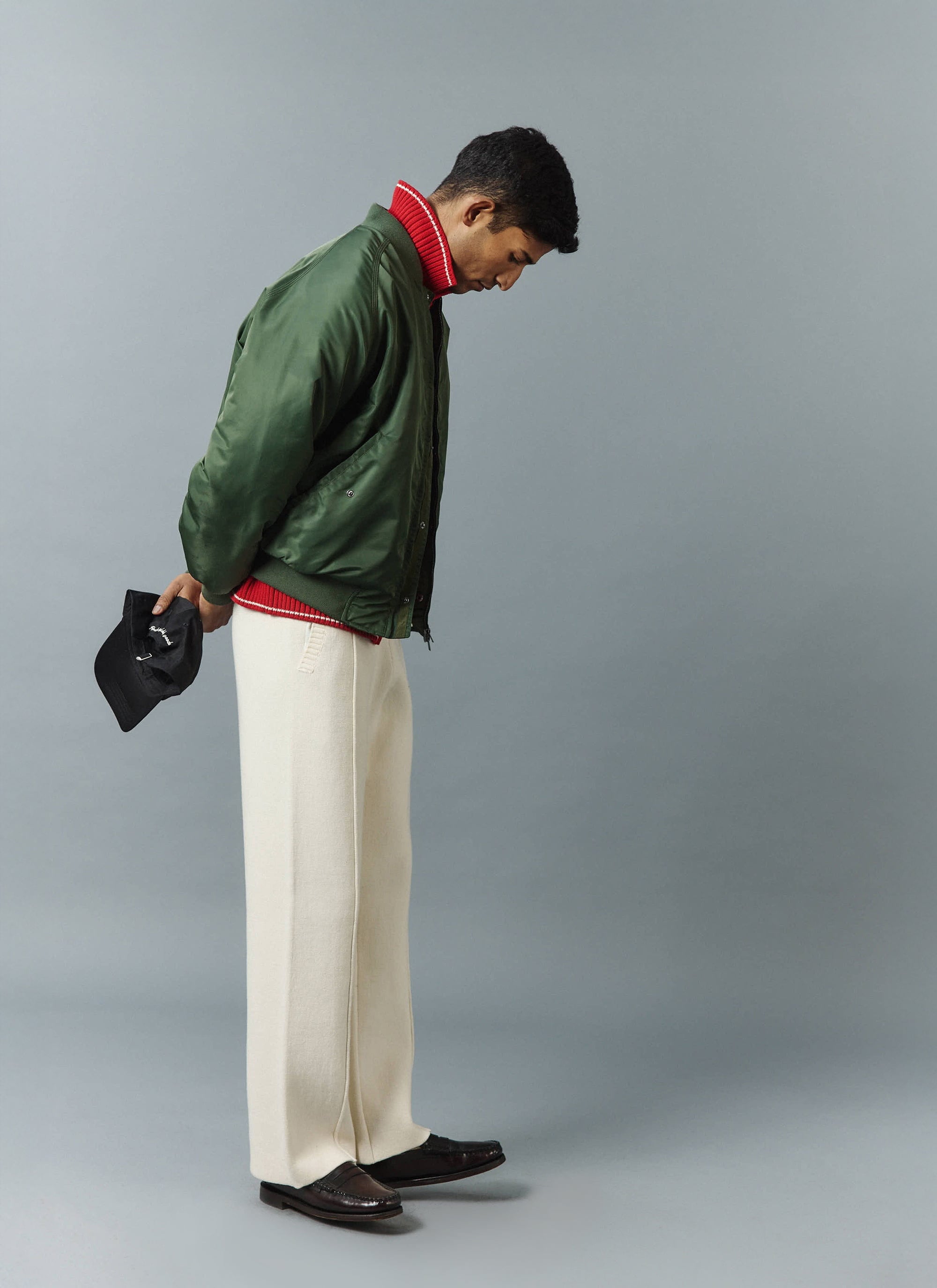 A model, shot from the side, wears the Percival Nylon Bomber Jacket in Forest over a red knit and cream trousers.