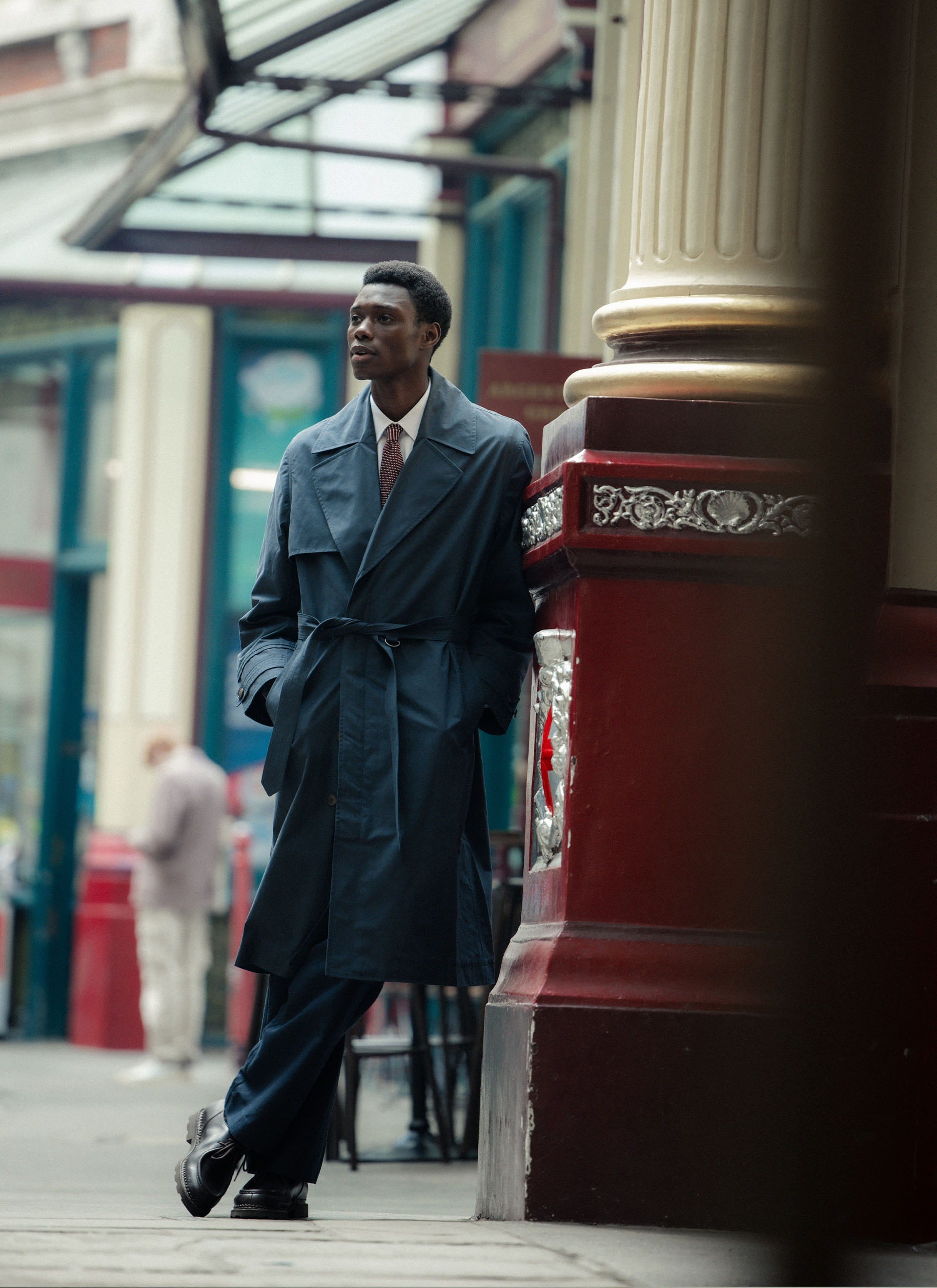 Navy cotton Percival x Front Office Mac Trench Coat, worn by a model leaning against an ornate pillar in the city.