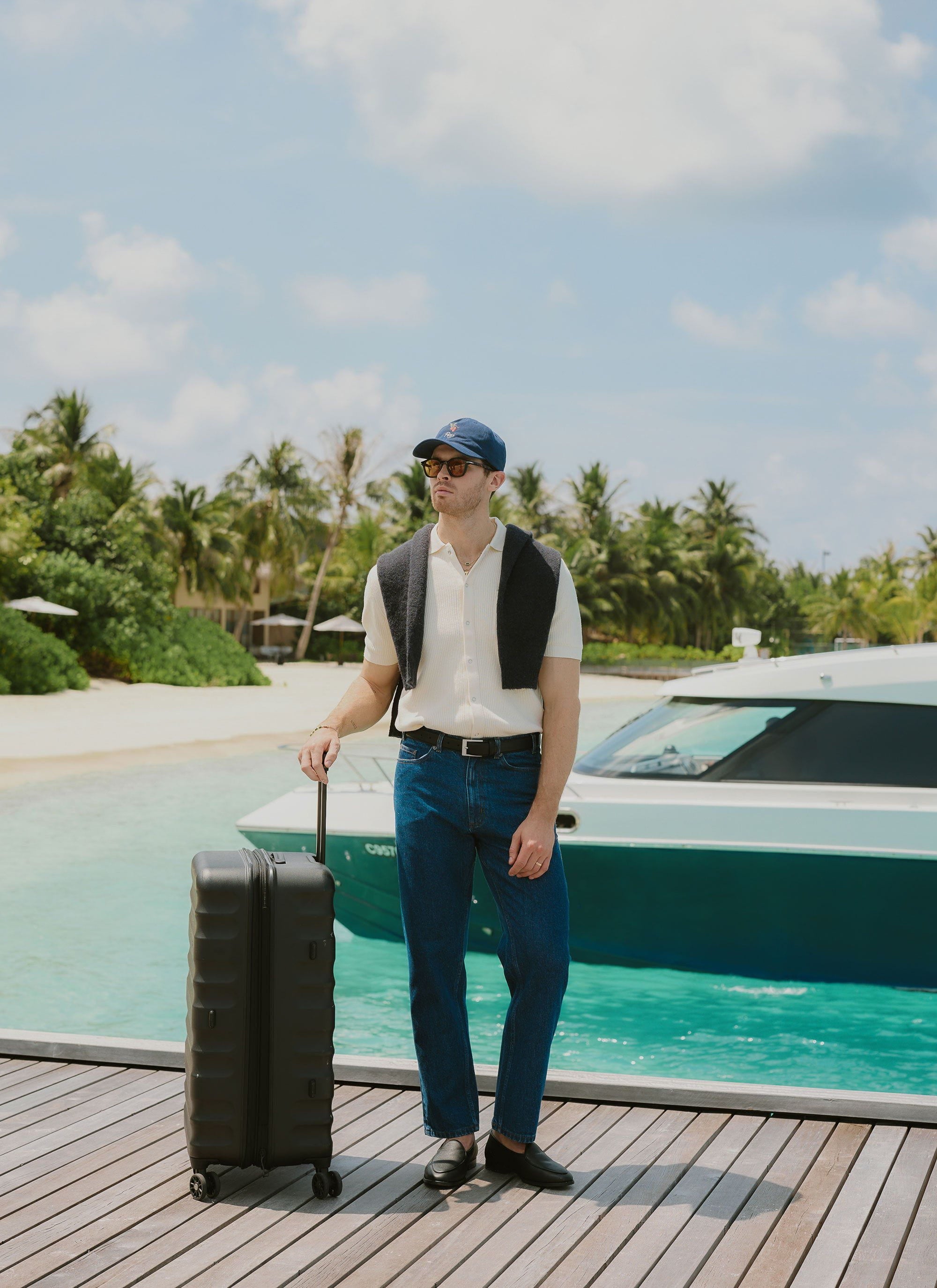 A man wears the Percival Pablo Cuban Shirt in cream, standing on a wooden pier with a suitcase next to a boat.