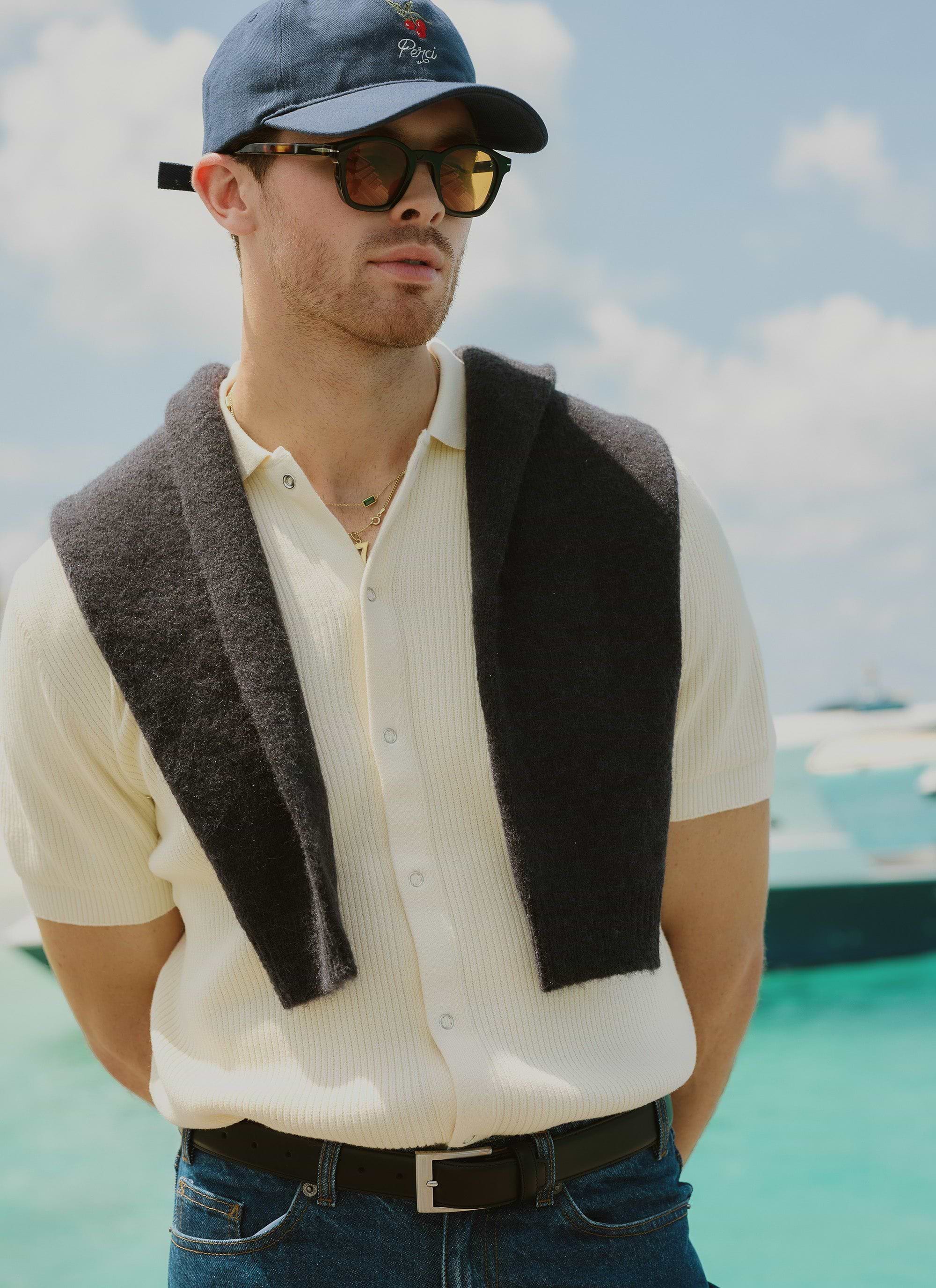 A model wears the Percival Pablo Cuban Shirt in cream, with a dark jumper draped over his shoulders, sunglasses and a cap.