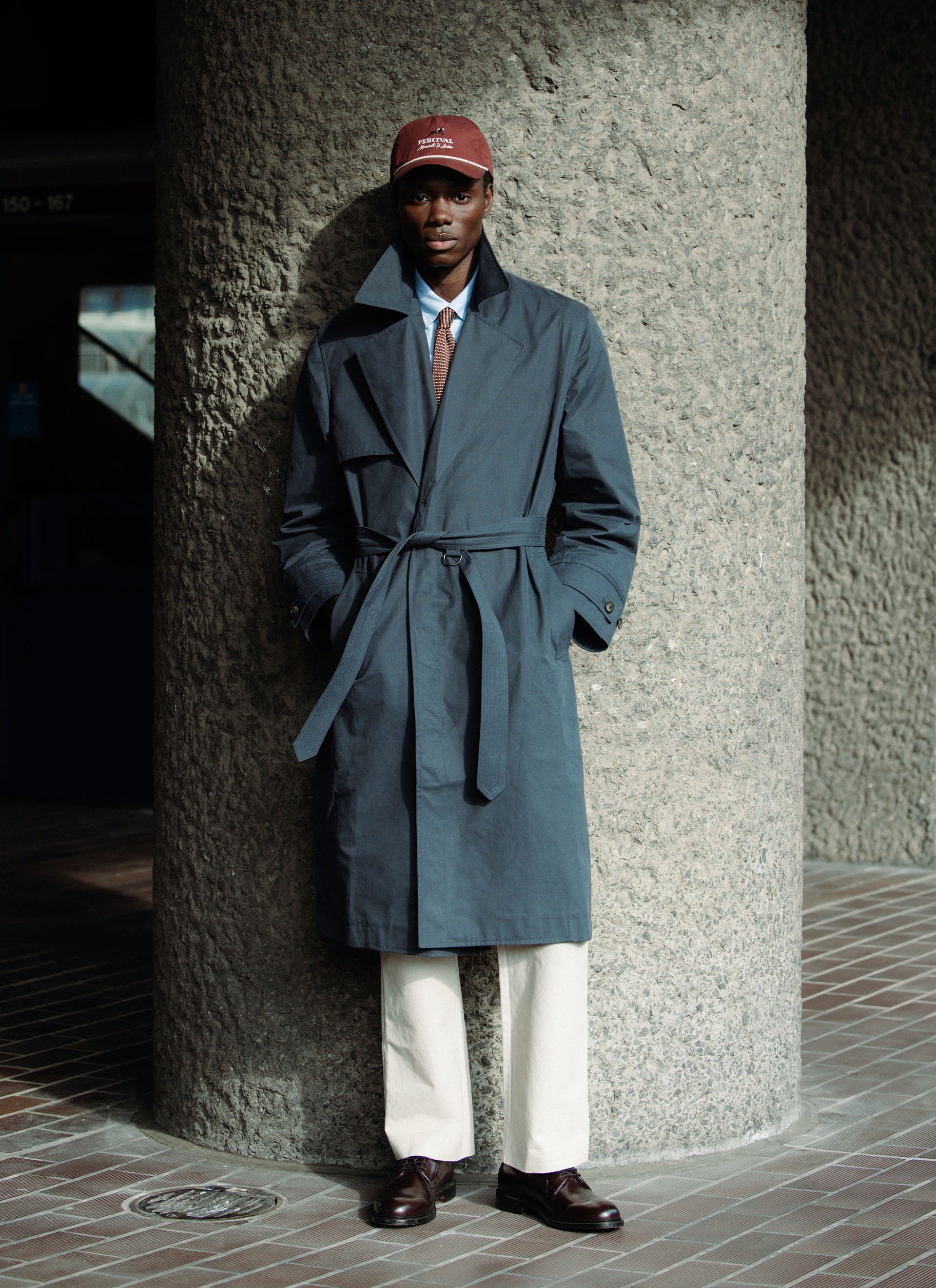 Navy Percival x Front Office Mac Coat, a belted trench style in peached cotton, worn by a model in an urban setting.