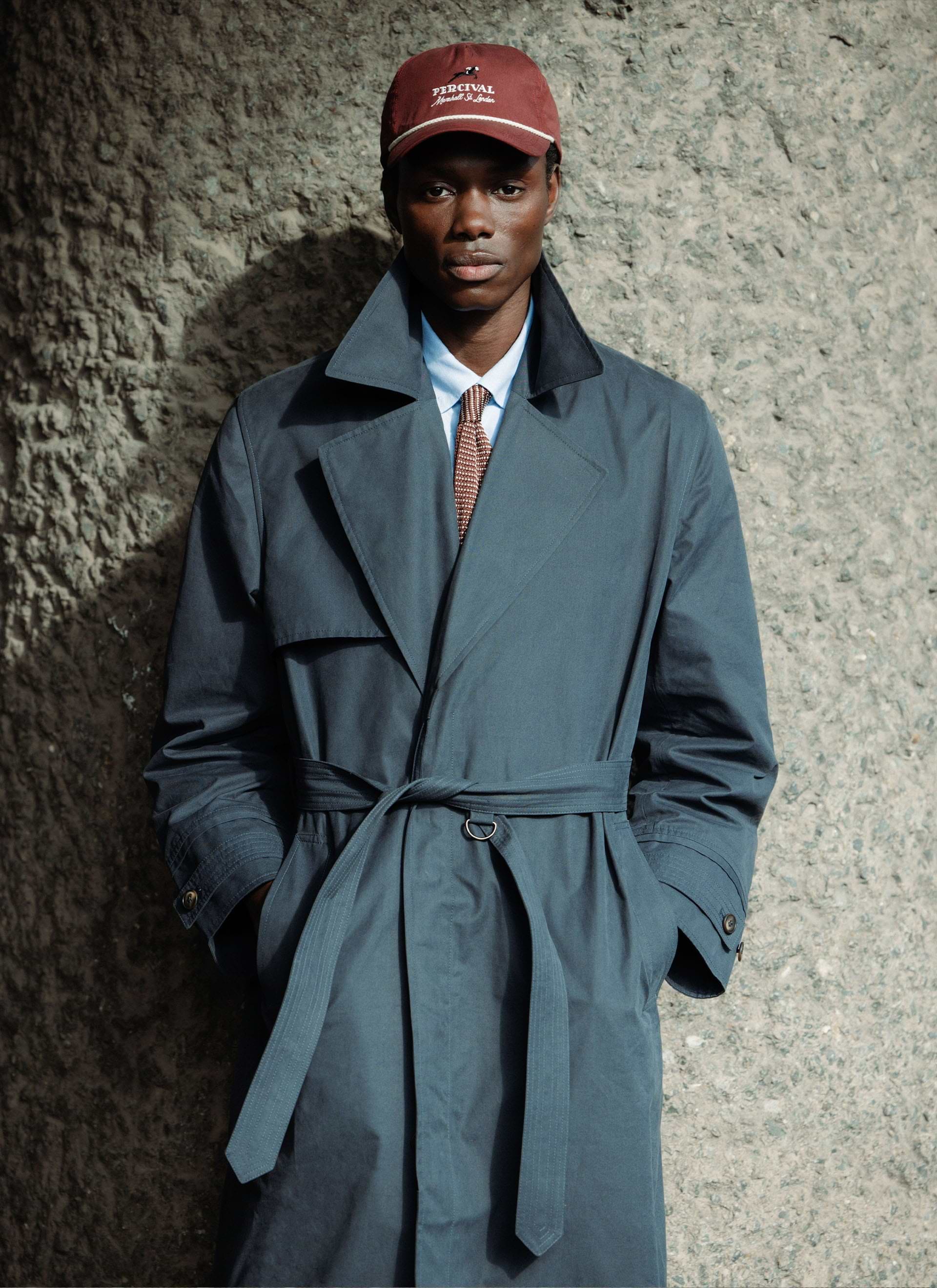 Navy cotton Mac Coat by Percival with a belted waist, worn by a model in a maroon cap against a concrete wall.