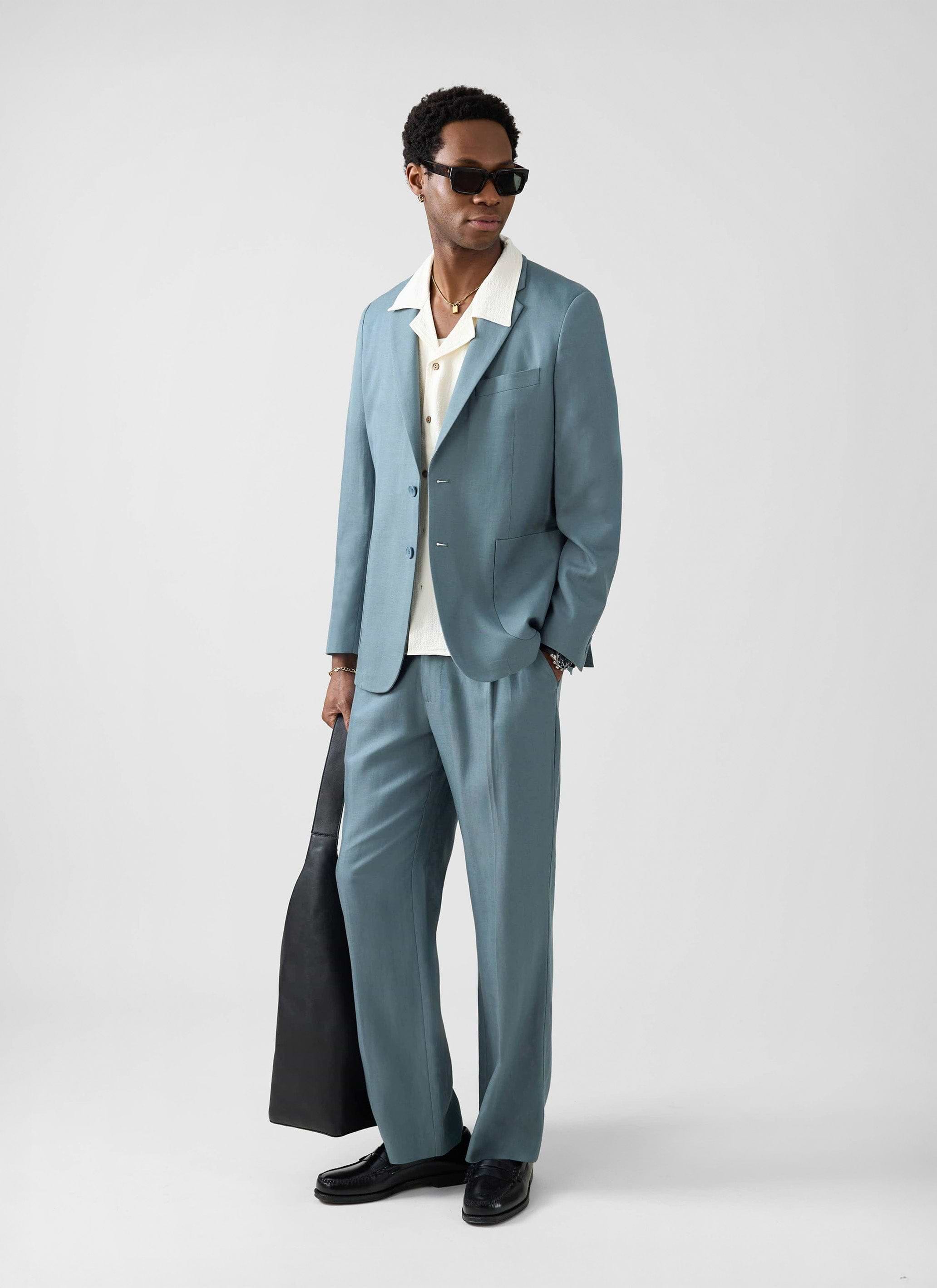 A man models the Percival Lyocell Regular Blazer and Trousers in Slate Blue, styled with sunglasses and a black bag.