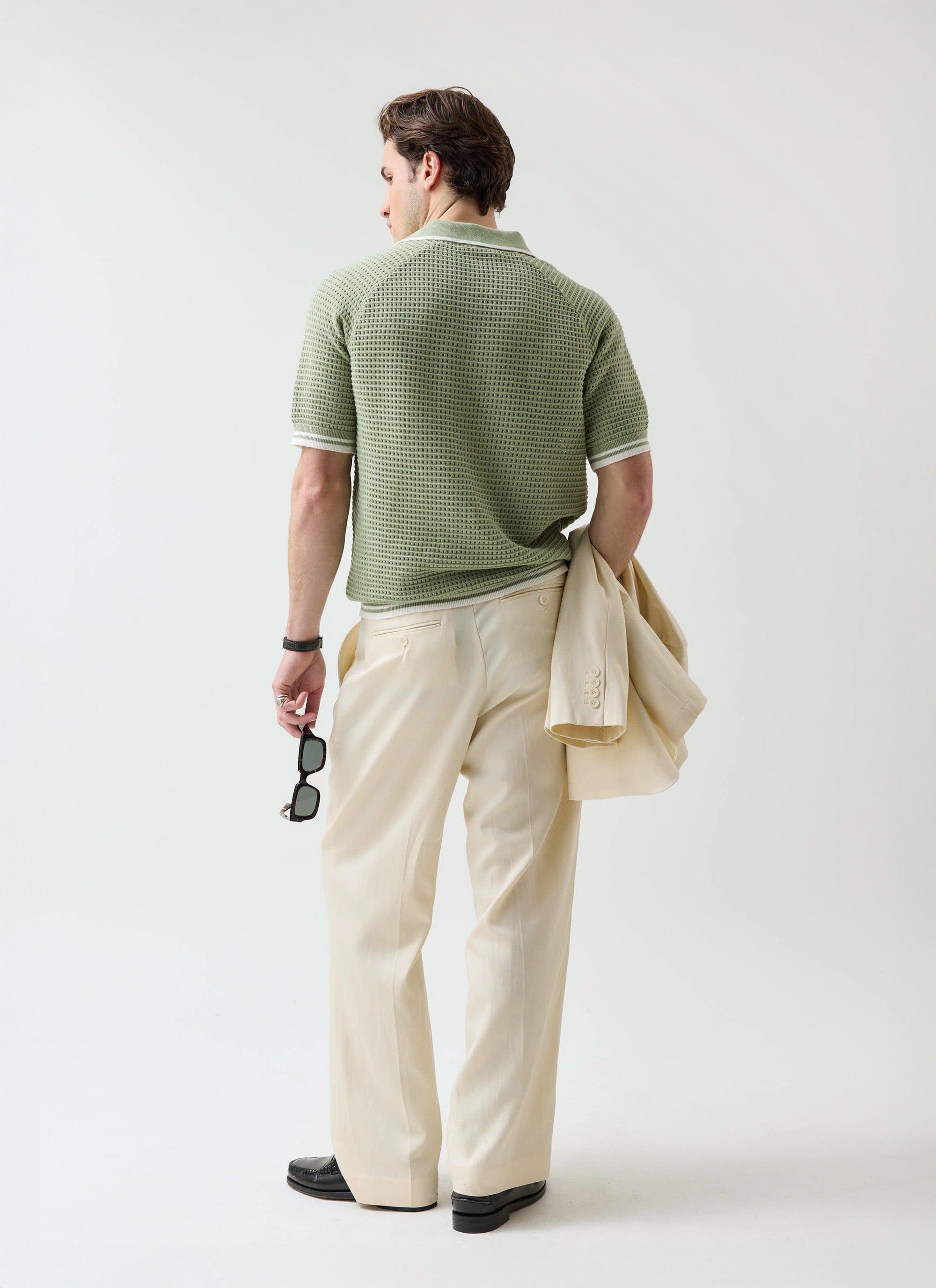 A model wearing the Percival Tennis Boxy Knitted Polo in sage with cream trousers and matching jacket, seen from behind.