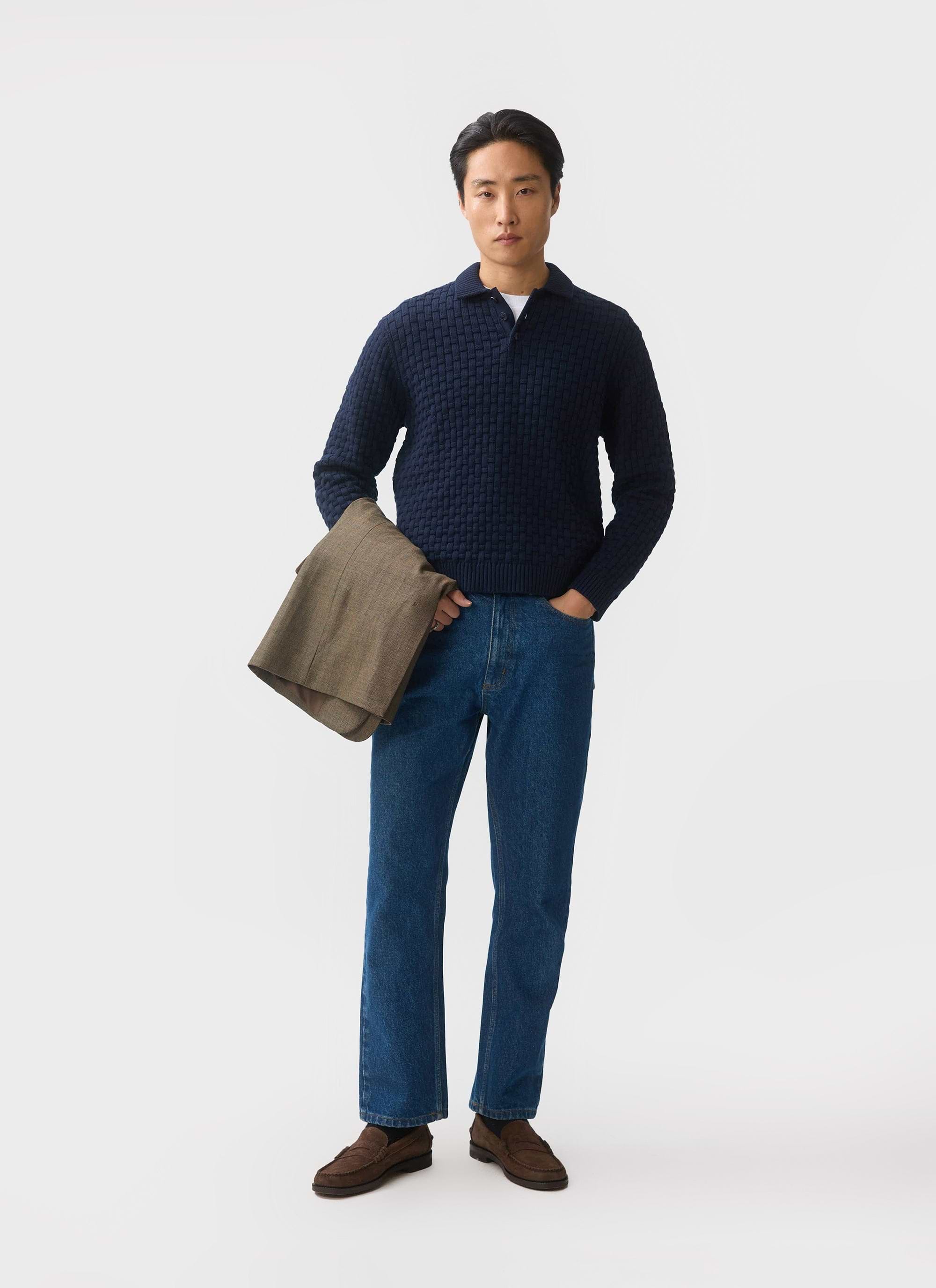 A model wears the Percival Archer Basketweave Knitted Shirt in navy, styled with blue jeans and holding a brown jacket.