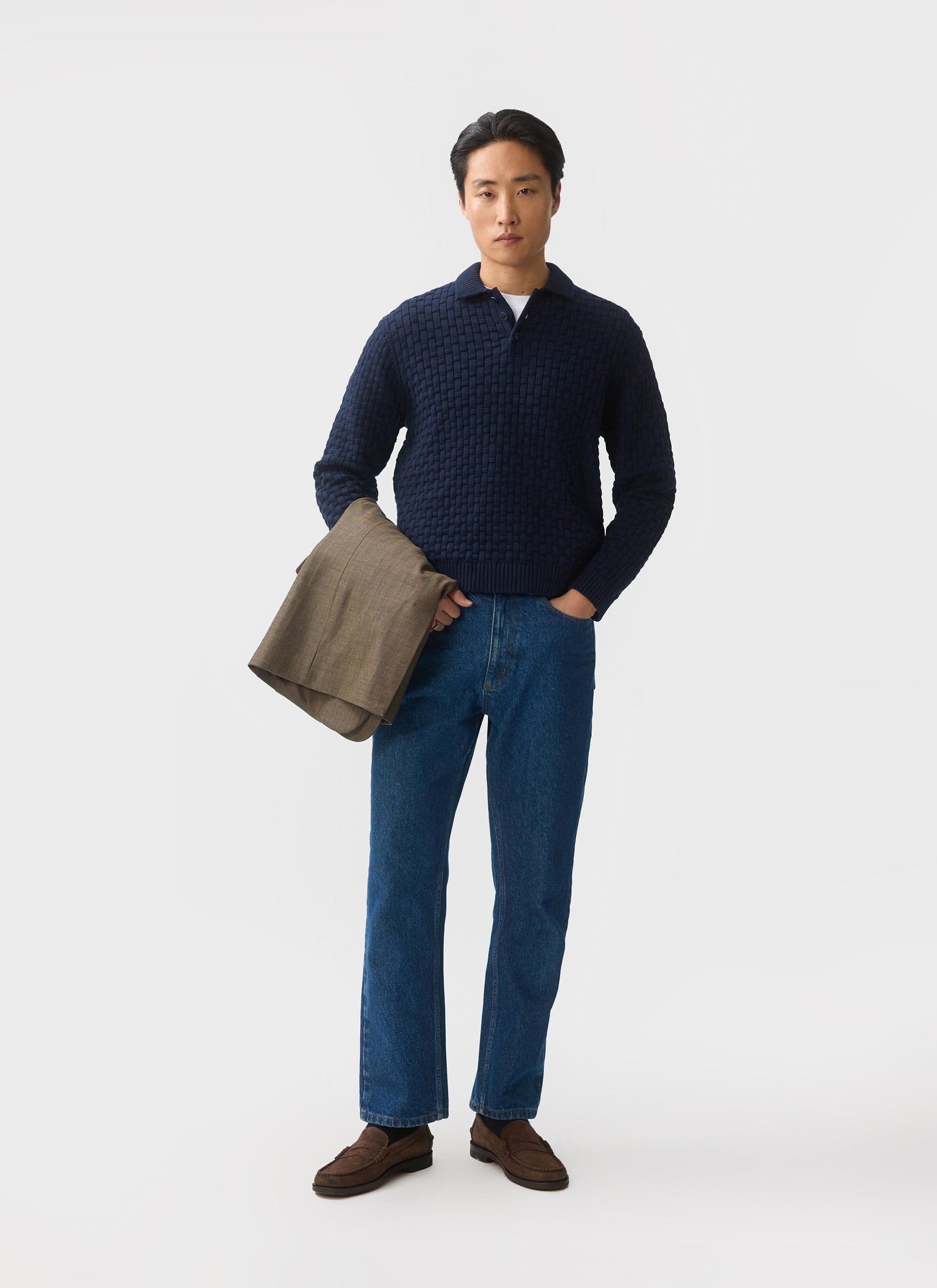 A model wears the Percival Archer Basketweave Knitted Shirt in navy, styled with blue jeans and holding a brown jacket.