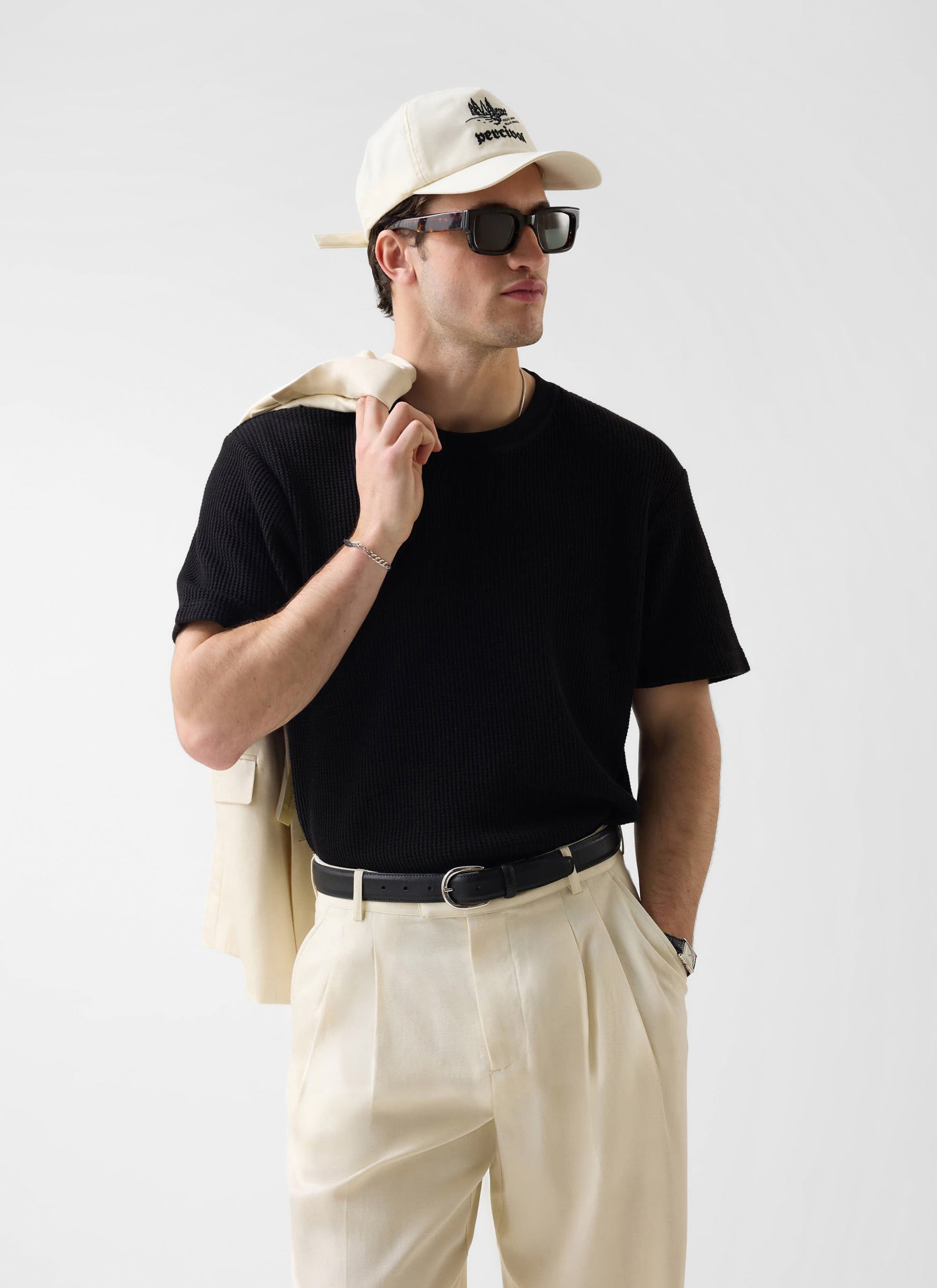 A model wears the Percival black Waffle T-Shirt, styled with cream pleated trousers, a matching jacket, and a Percival cap.