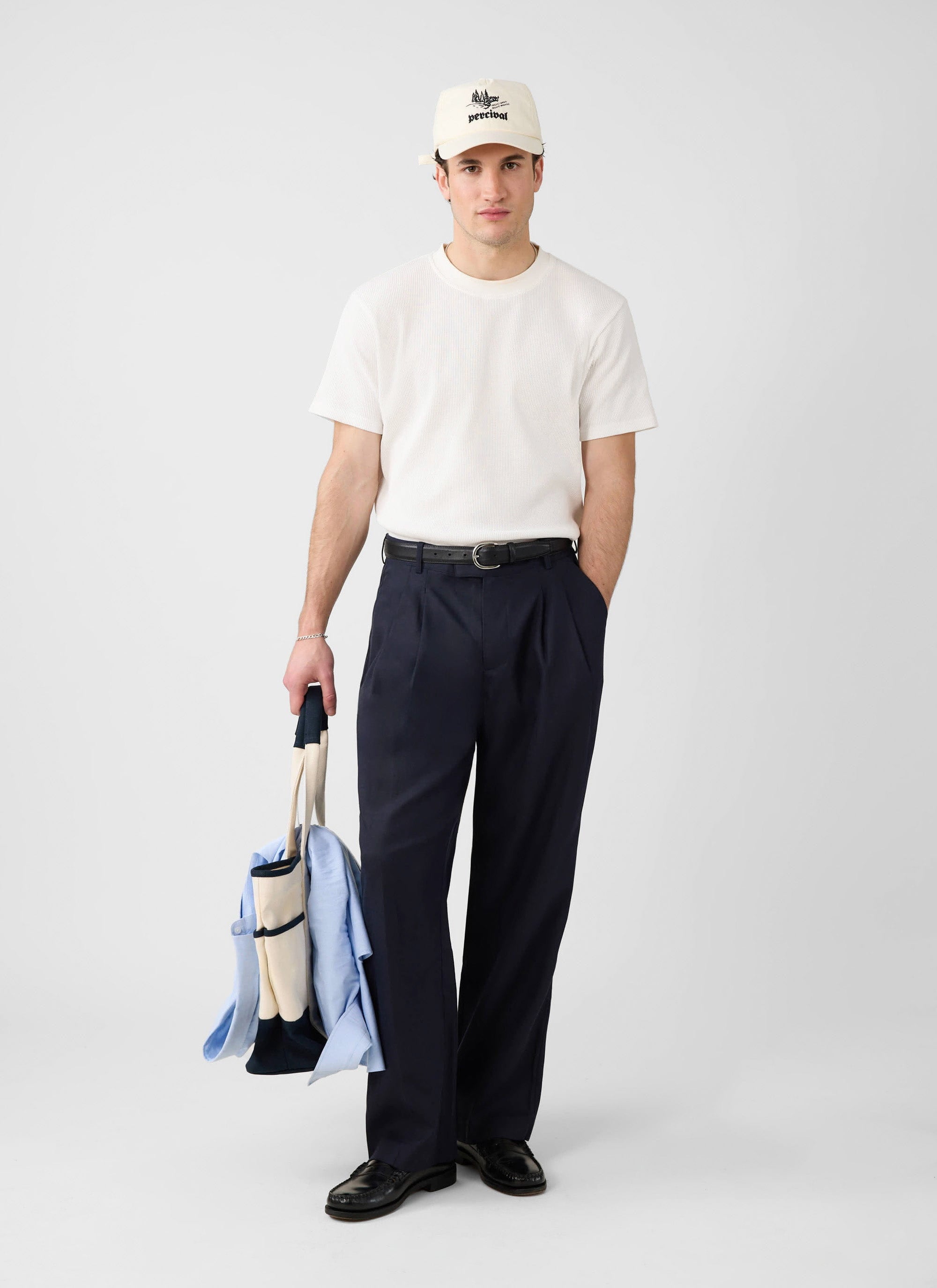 A model wears the Percival Waffle Short Sleeve T-Shirt in white, paired with navy trousers, a cap, and holding a tote bag.