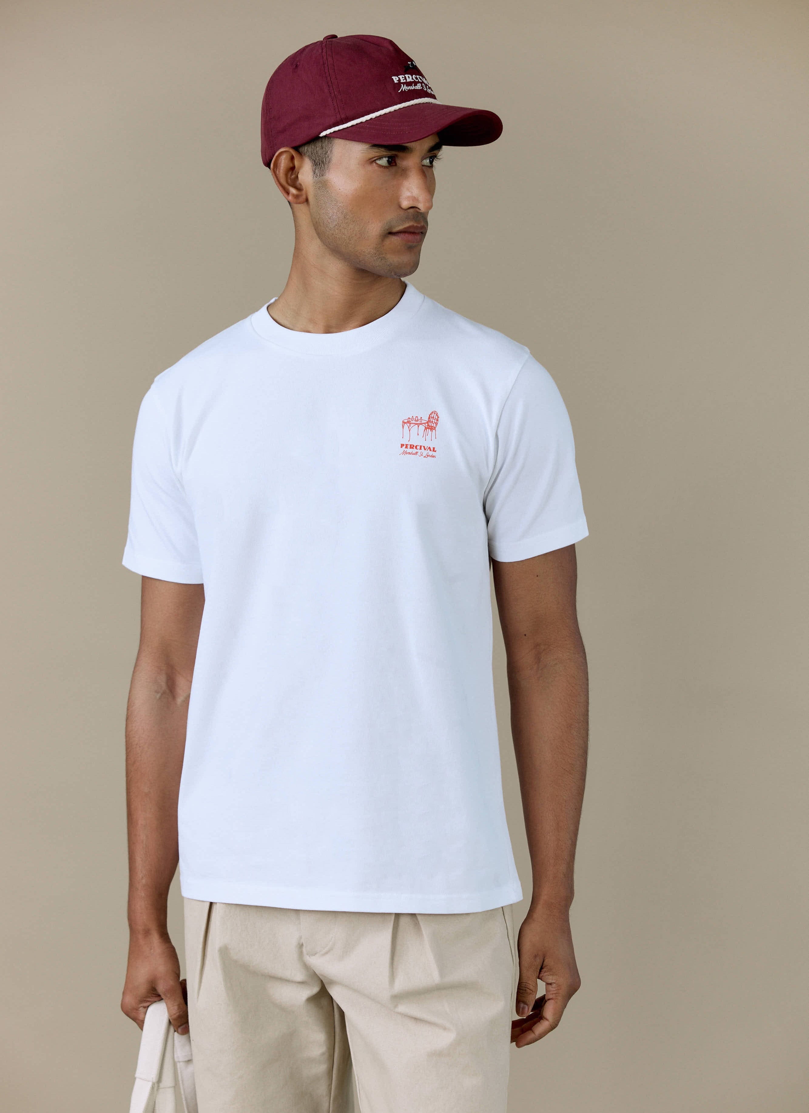 A model wears the white Perci Fine Wines T-shirt from Percival, featuring a small red chest motif and burgundy cap.