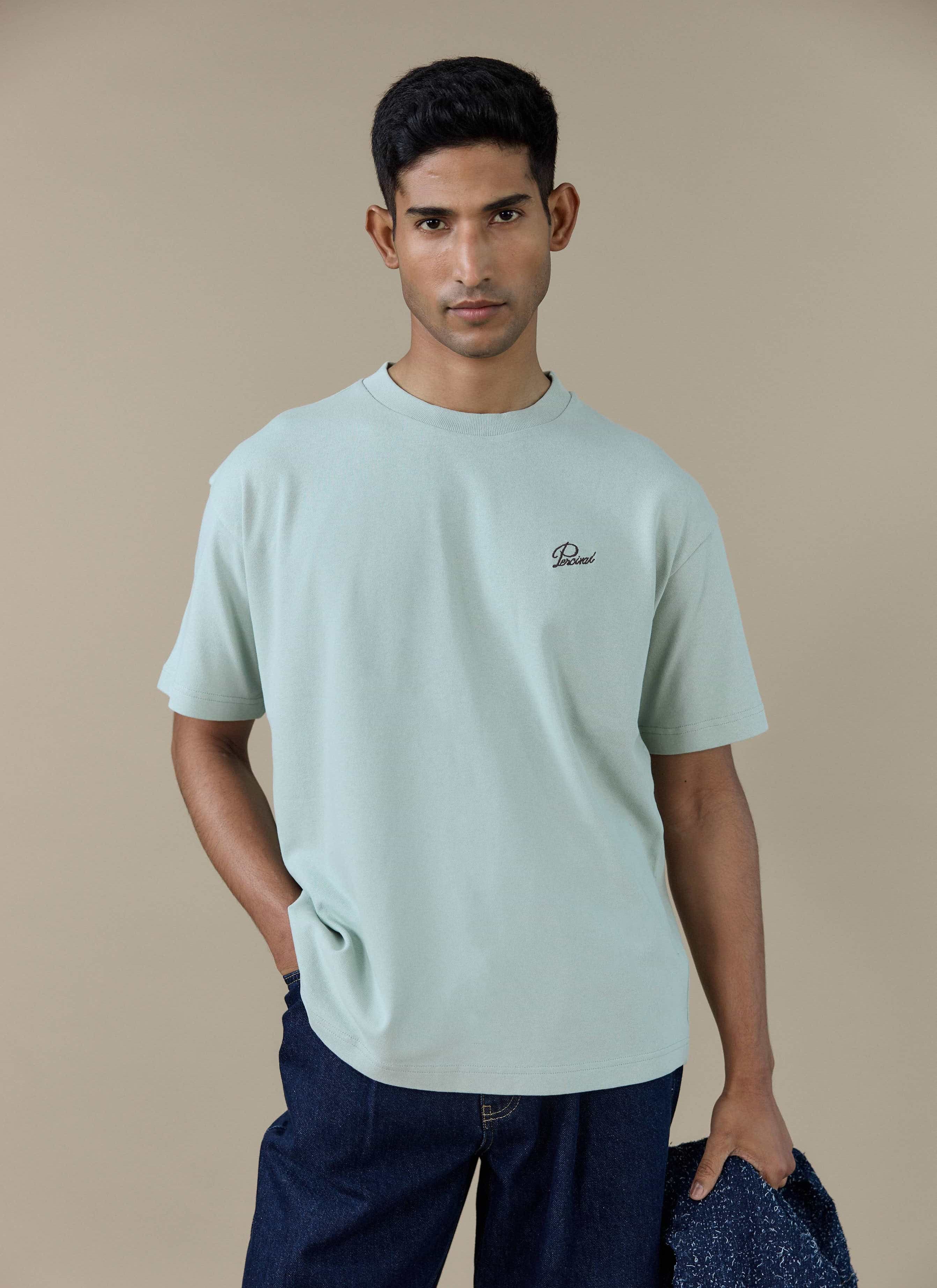 A model wearing the Percival Perci Script T Shirt in sage green, showing the script embroidery on the chest.