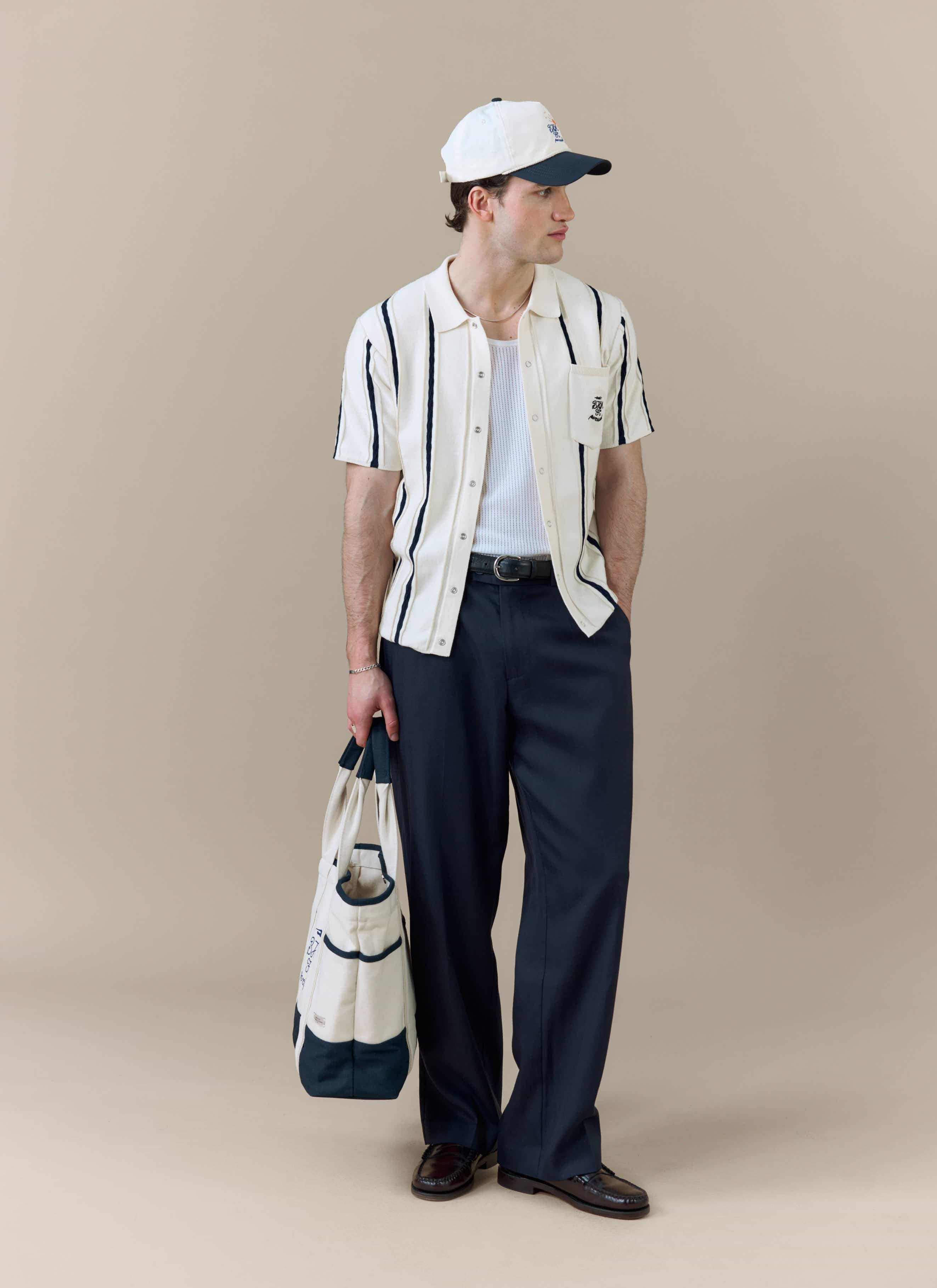 A model wears the Percival x Brighton & Hove Albion F.C. Cable Stripe Knitted Shirt with dark trousers, a cap and a tote bag.