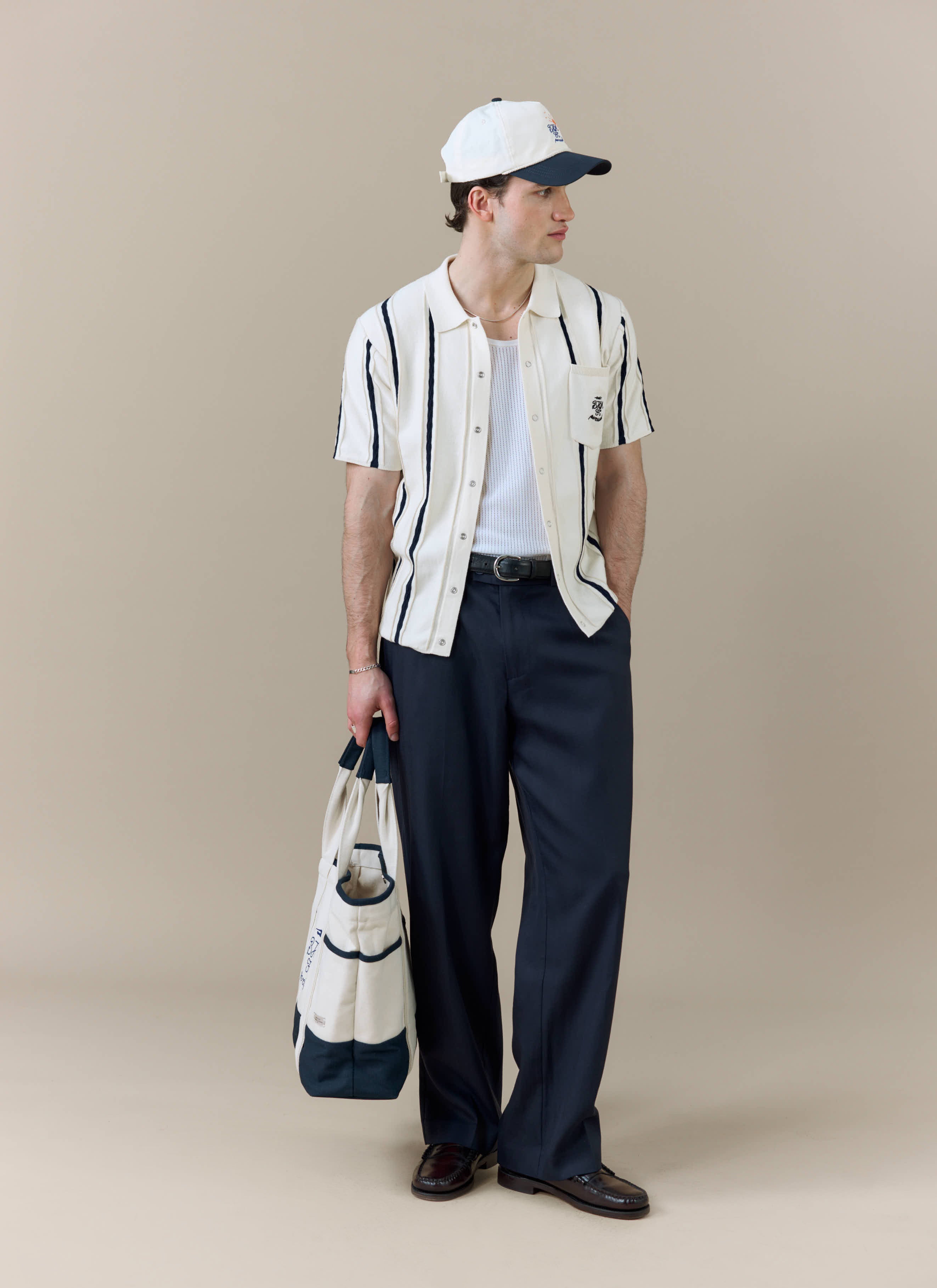 A model wears the Percival x Brighton & Hove Albion F.C. Cable Stripe Knitted Shirt with dark trousers, a cap and a tote bag.