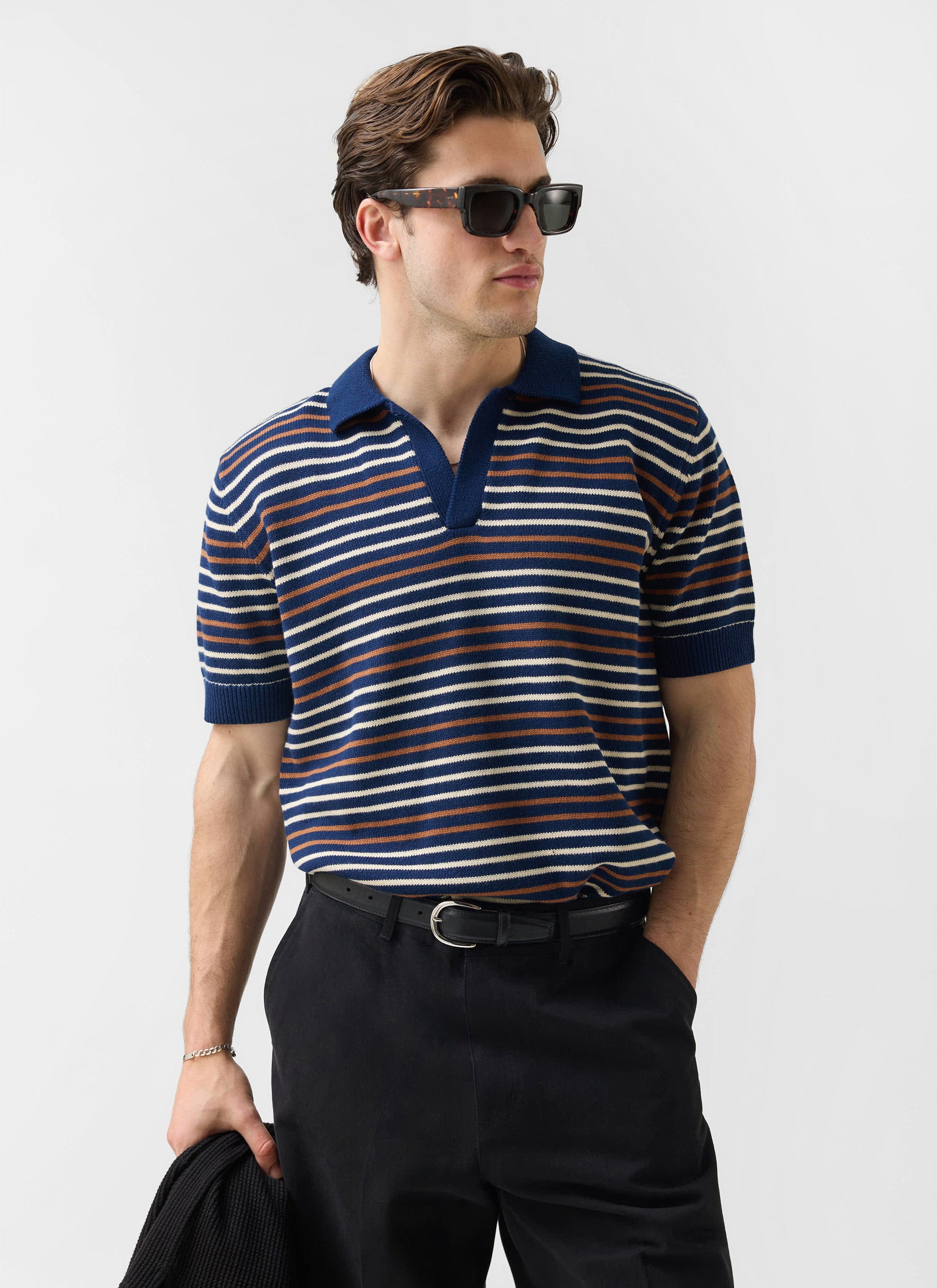 A man models the navy striped Douglas Boxy Knitted Polo from Percival, wearing sunglasses and black trousers.