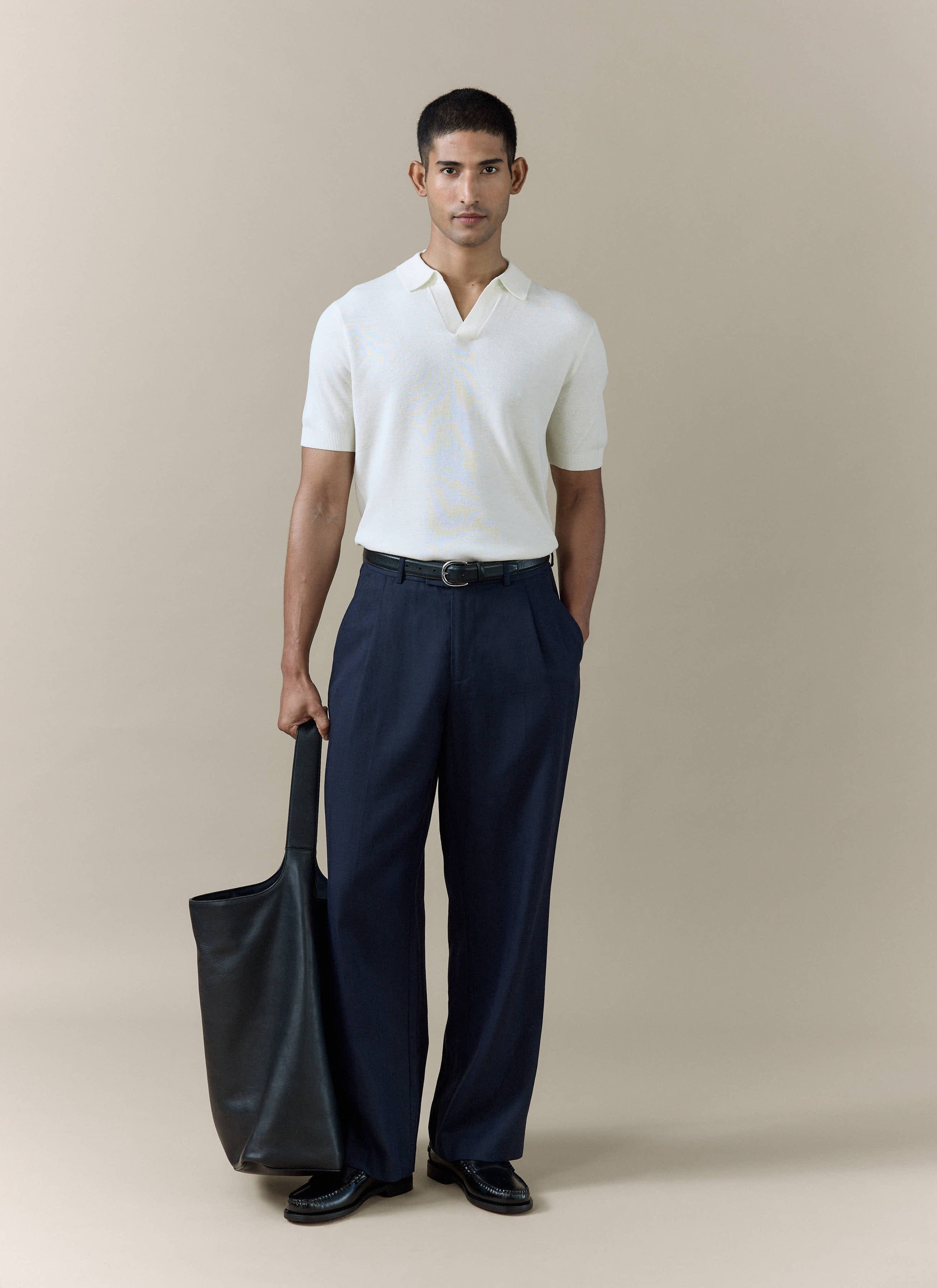 Percival Laurence cream knitted polo in a linen-lyocell blend, styled with navy pleated trousers and a black tote bag.