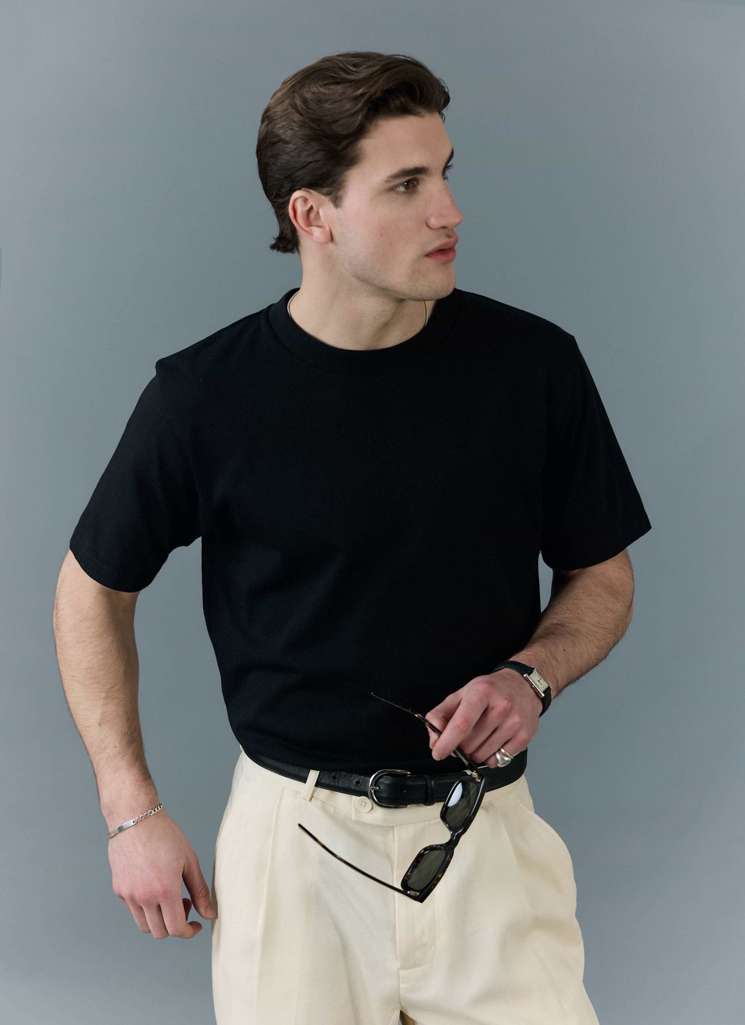A man models the Percival black organic cotton t-shirt, styled with cream trousers and sunglasses.