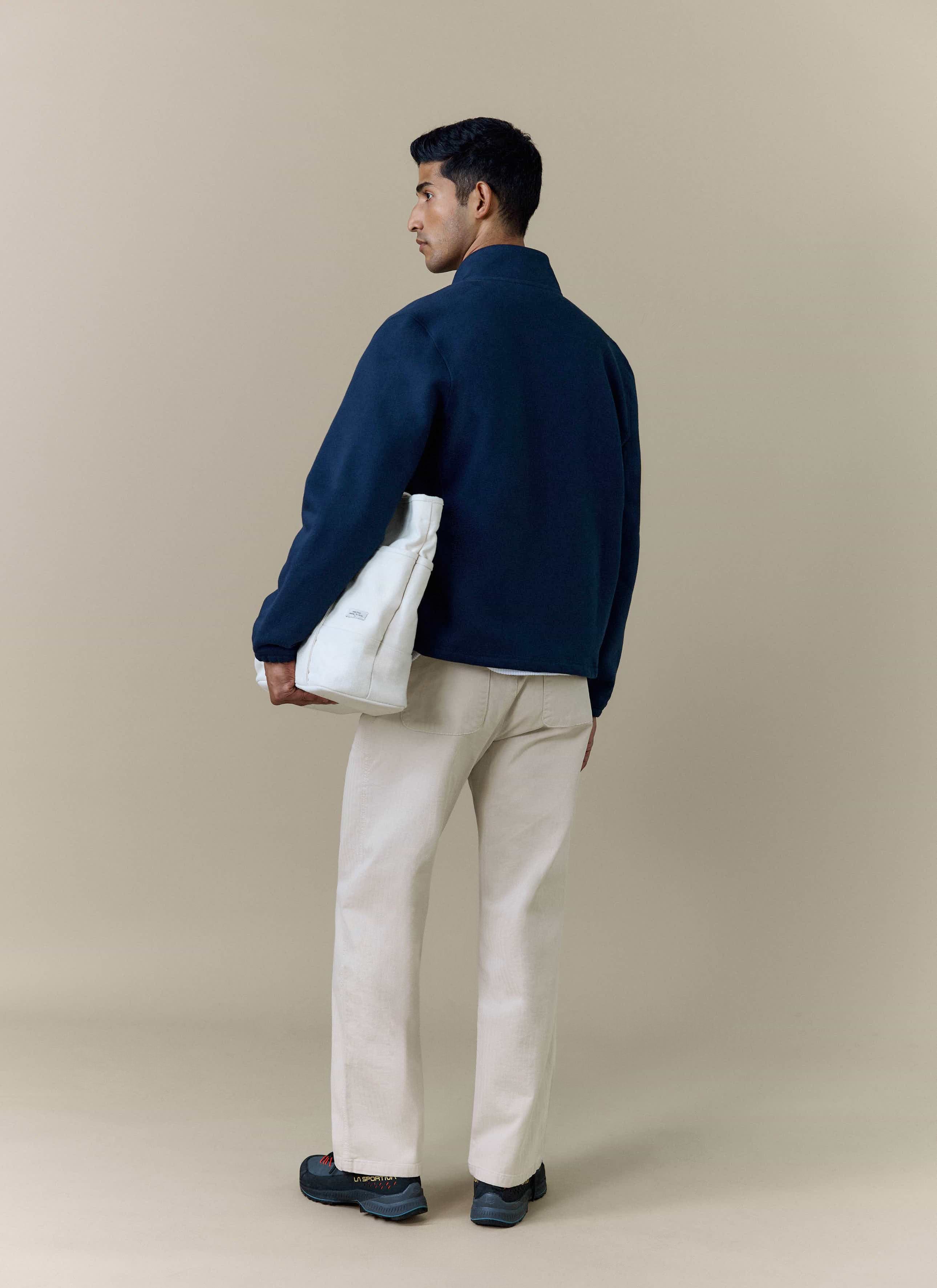 A model seen from behind wearing the Percival Curtis Boxy Harrington in navy, styled with cream trousers and a tote bag.