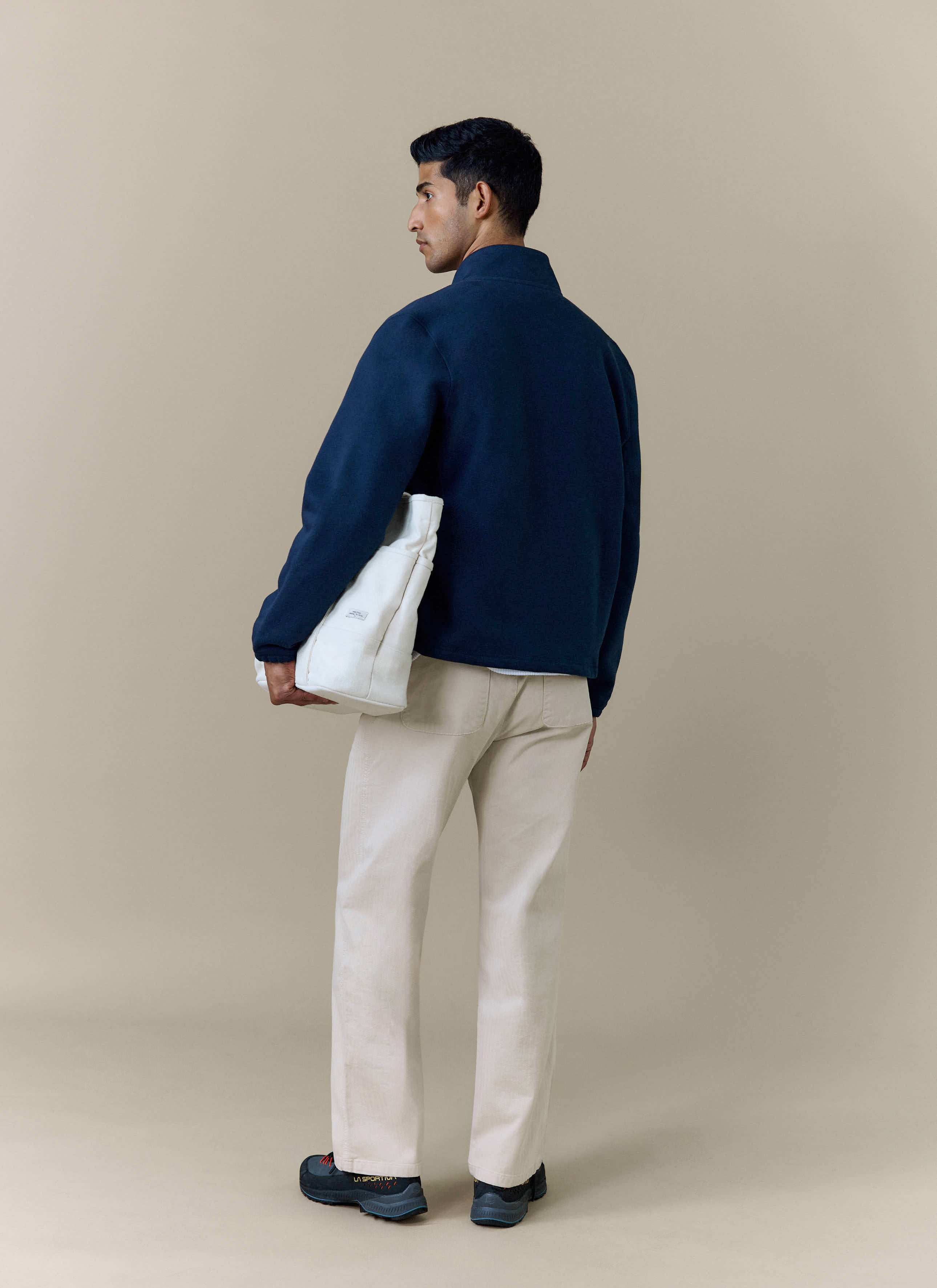 A model seen from behind wearing the Percival Curtis Boxy Harrington in navy, styled with cream trousers and a tote bag.