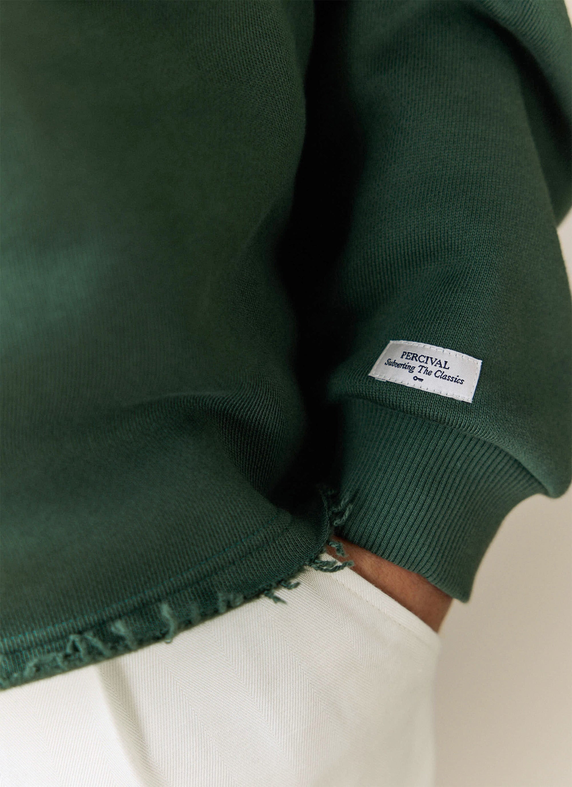 Close-up of the Percival Dudley Boxy Sweatshirt in forest green, showing the ribbed cuff and woven brand label on the sleeve.