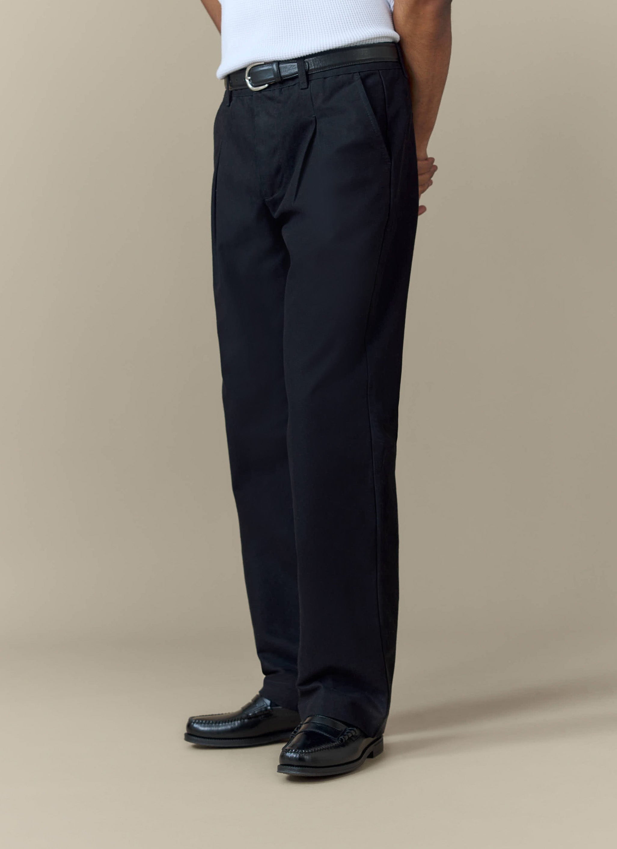 A model wears the black Seb Inverted Pleat Trousers from Percival, styled with a white vest and black loafers.