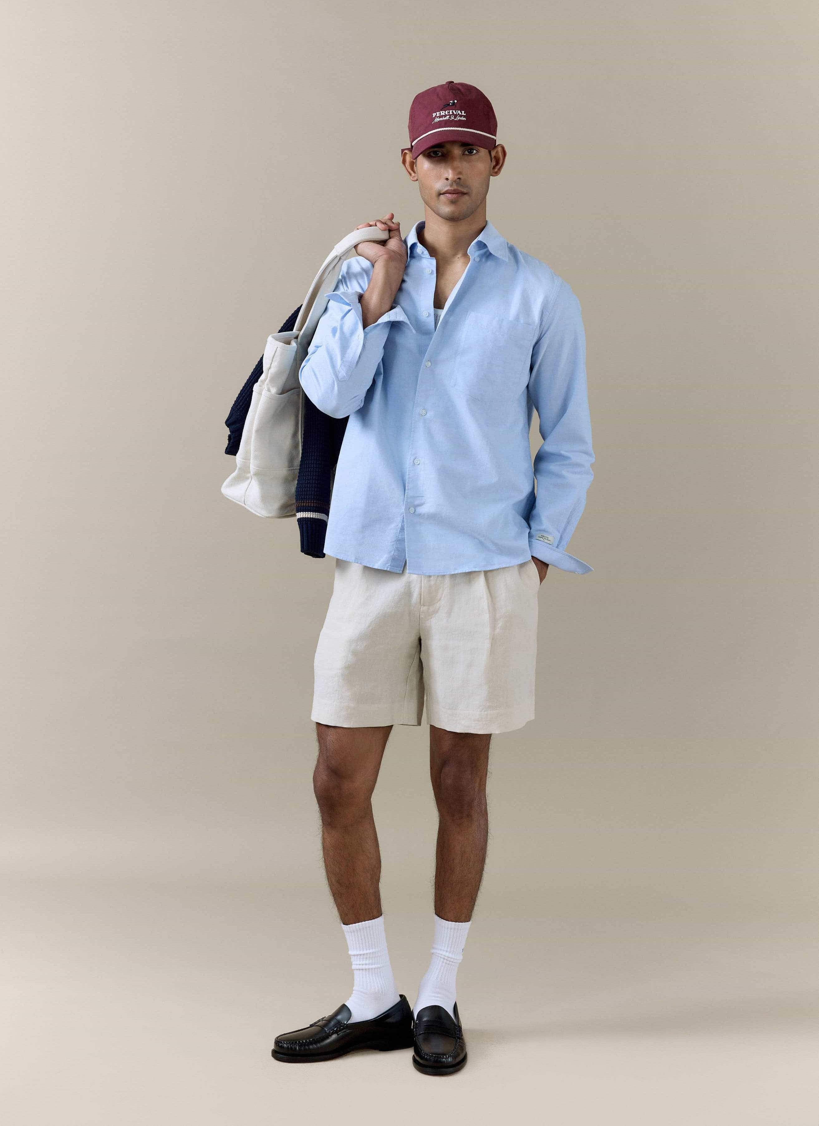 Cream Percival pleated linen shorts worn by a model with a light blue shirt, burgundy cap, and a white canvas tote bag.