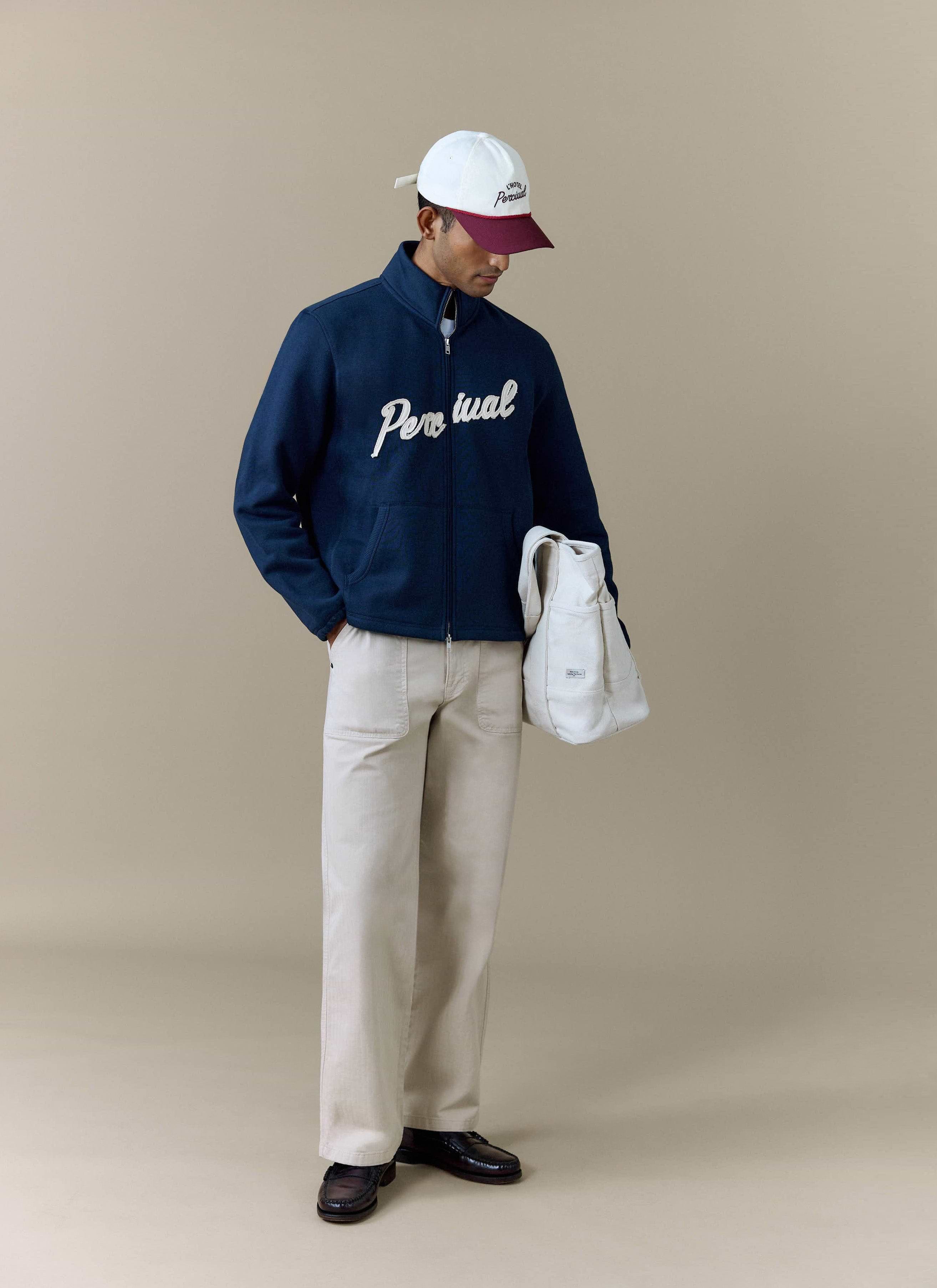 A model wears the navy Curtis Boxy Zip Funnel Neck Harrington from Percival, styled with beige trousers and a tote bag.