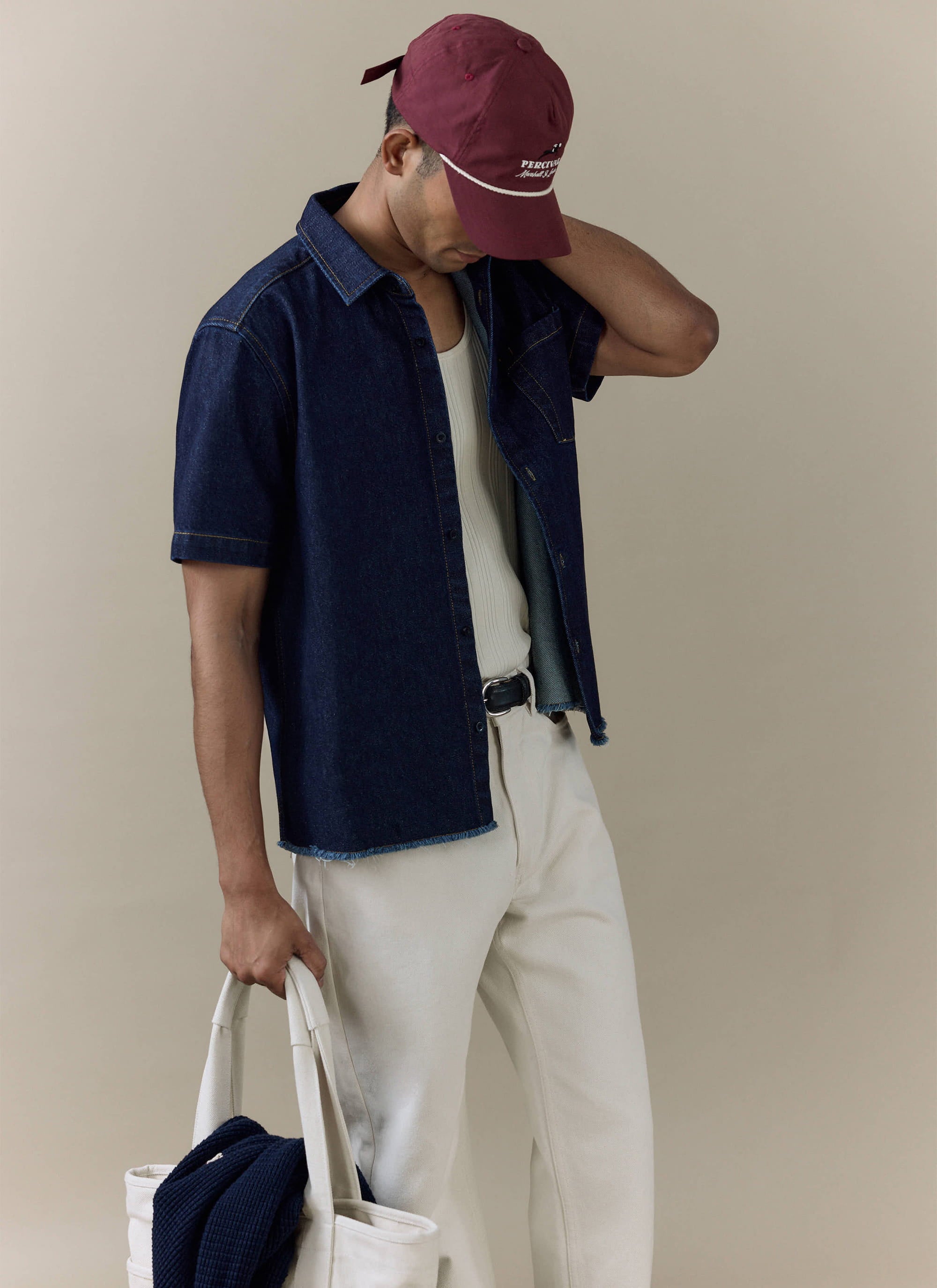 Model wearing the Percival Harley Denim Cropped Boxy Shirt in dark indigo, styled with cream trousers and a burgundy cap.