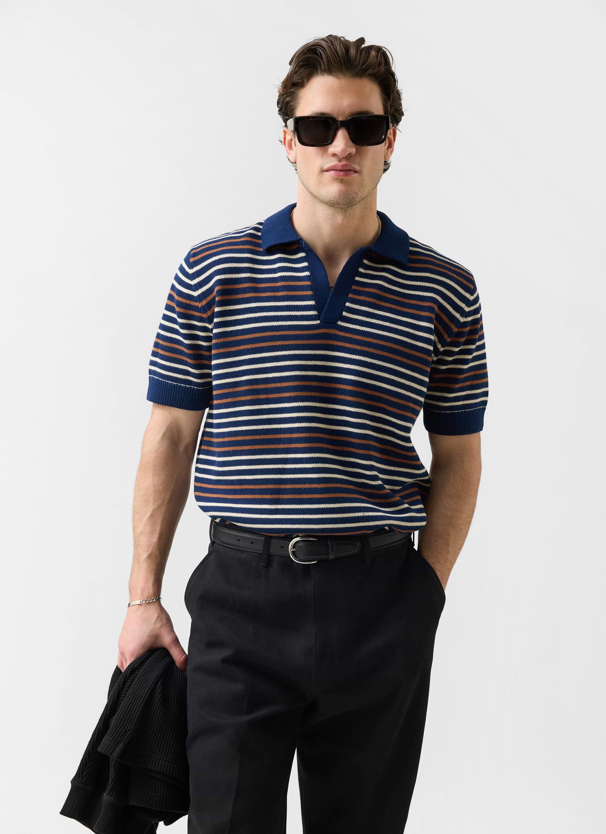 A model wears the navy striped Percival Douglas Boxy Knitted Polo, styled with smart black trousers and sunglasses.