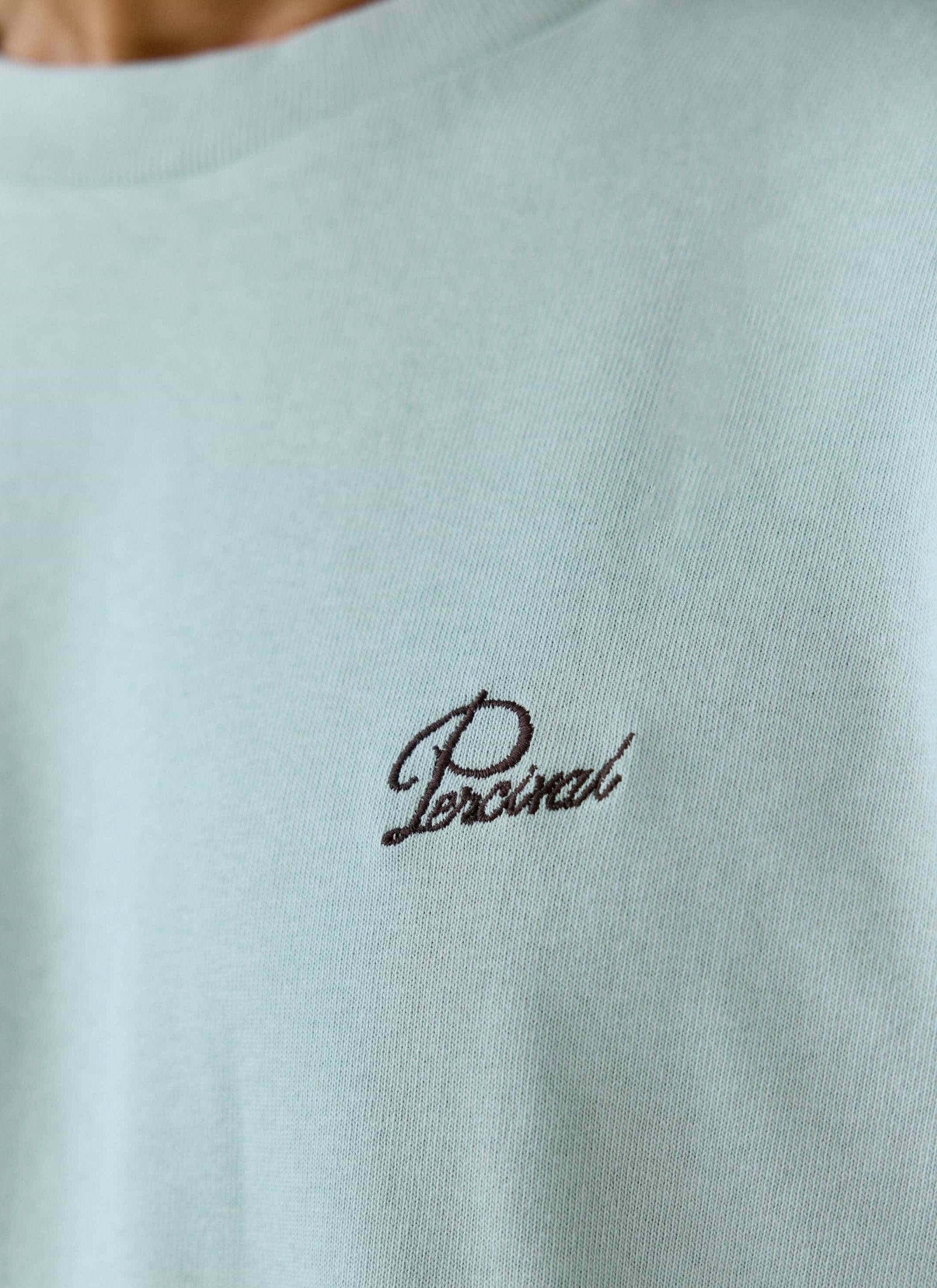 A close-up of the dark script embroidery on the chest of the sage green Perci Script T Shirt from Percival.