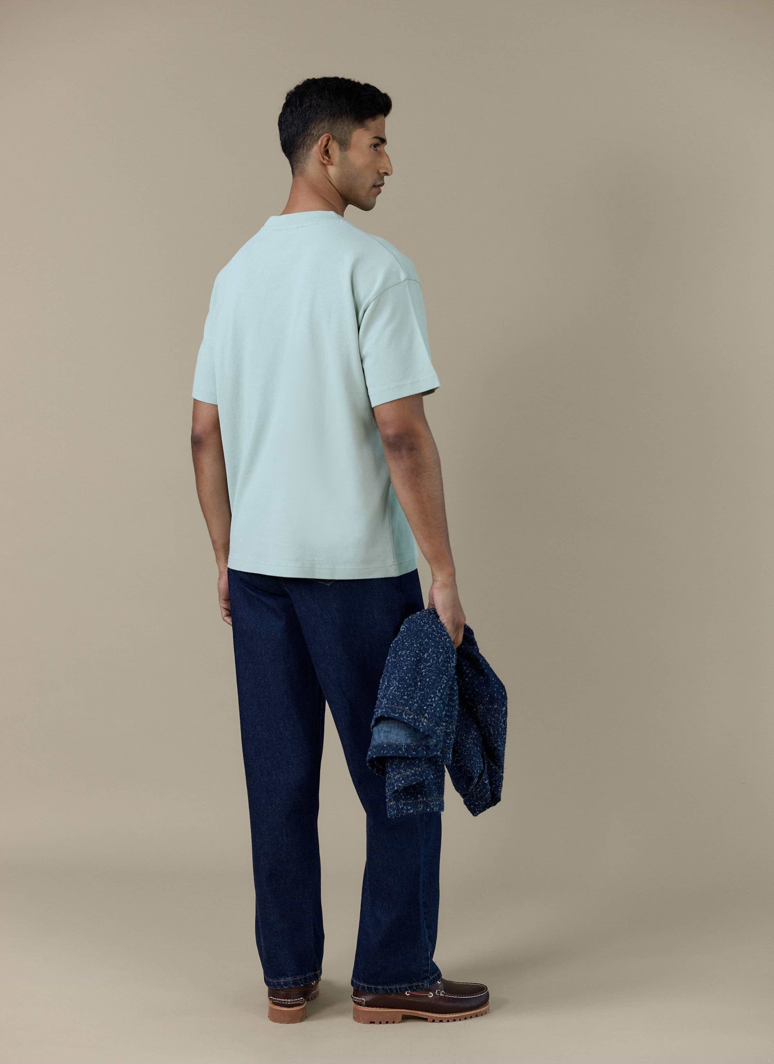Back view of a model wearing the Percival Perci Script T Shirt in sage, paired with dark blue trousers.