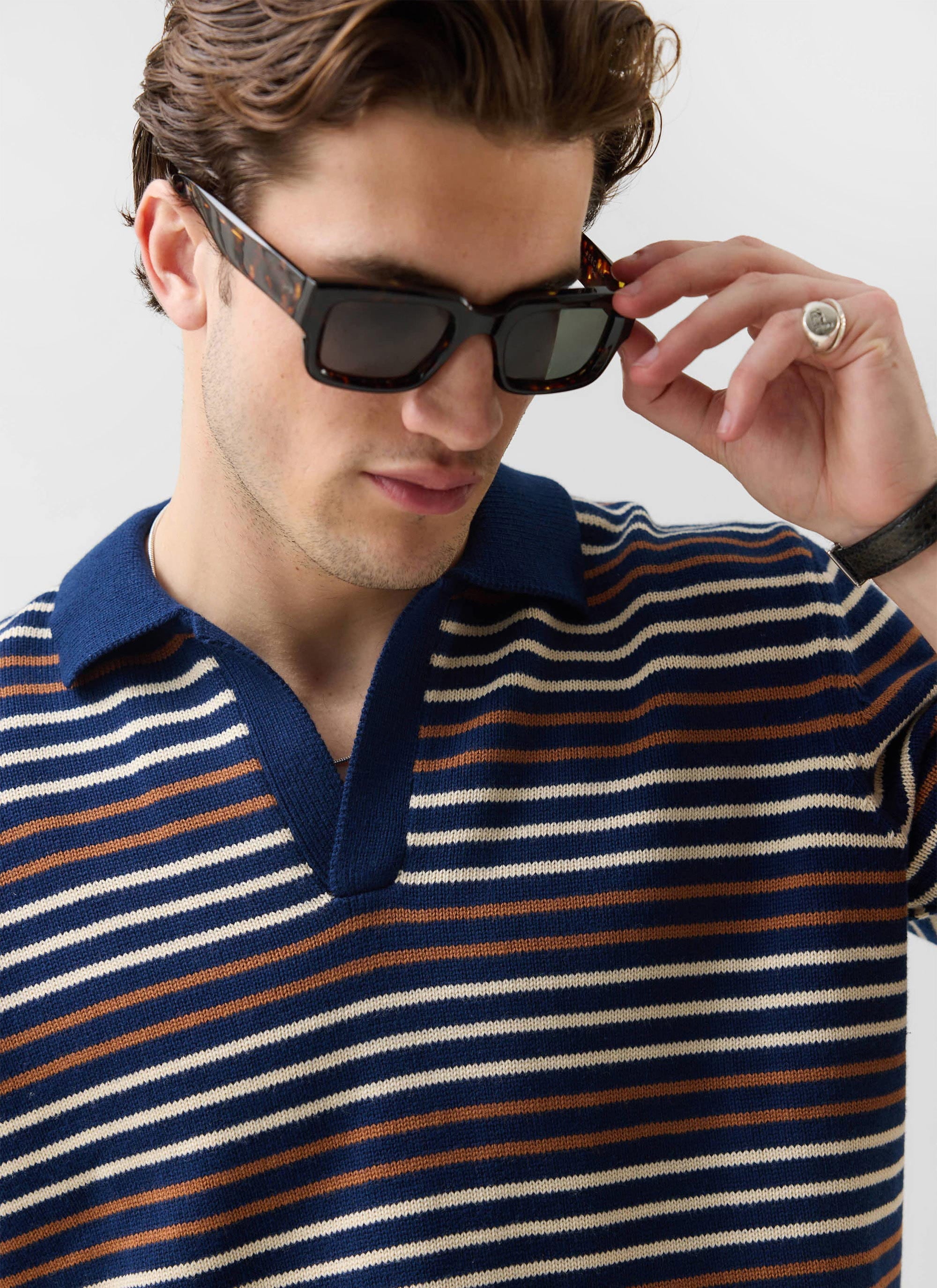 A close-up of a model wearing the Percival Douglas Boxy Knitted Polo in navy with stripes, adjusting his sunglasses.