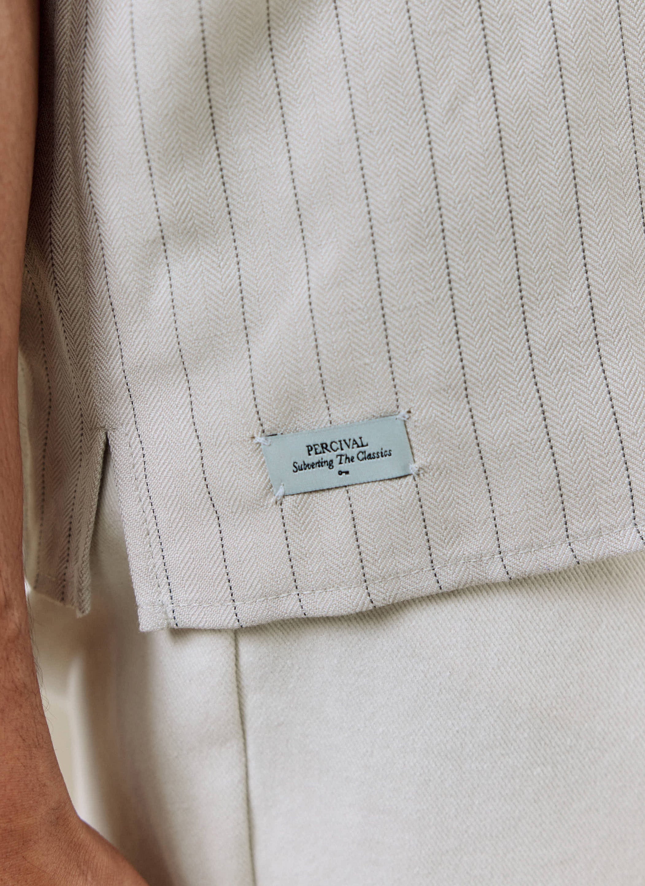 Close-up of the Percival cream Striped Boxy Shirt, showing the pinstripe herringbone weave, side split, and branded tag.