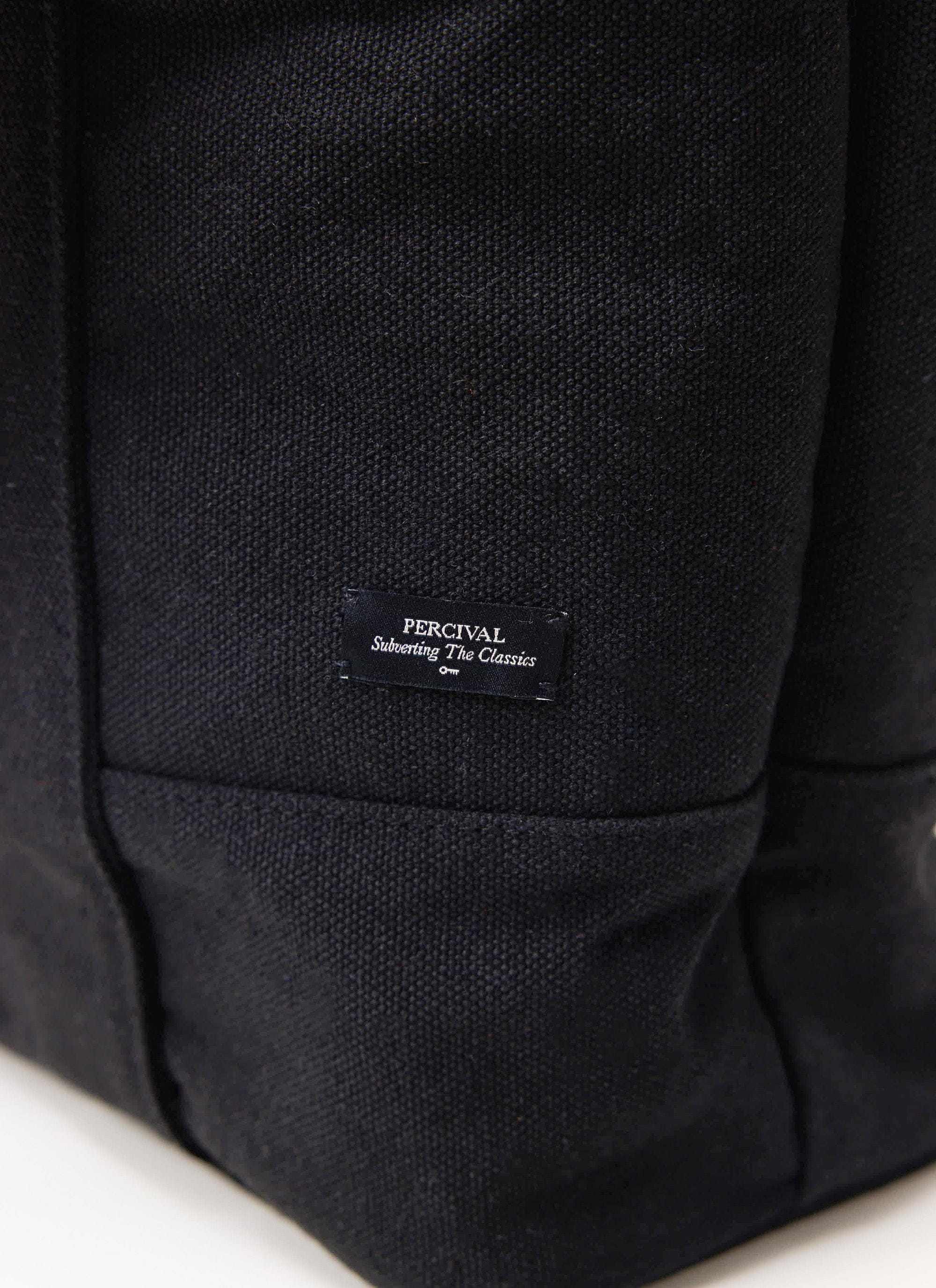 A close-up of the woven 'Percival Subverting The Classics' label on the brand's black canvas tote bag.