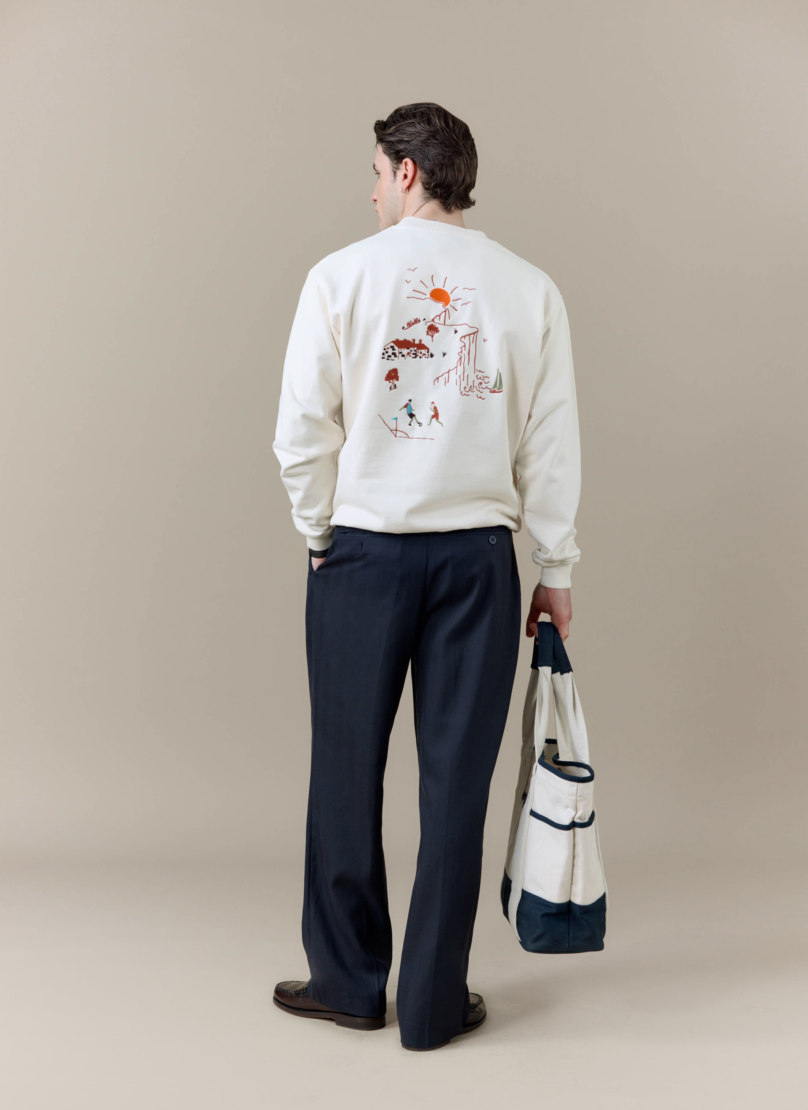 A model seen from behind wearing the Percival x Brighton F.C. cream sweatshirt, carrying the navy and cream tote bag.