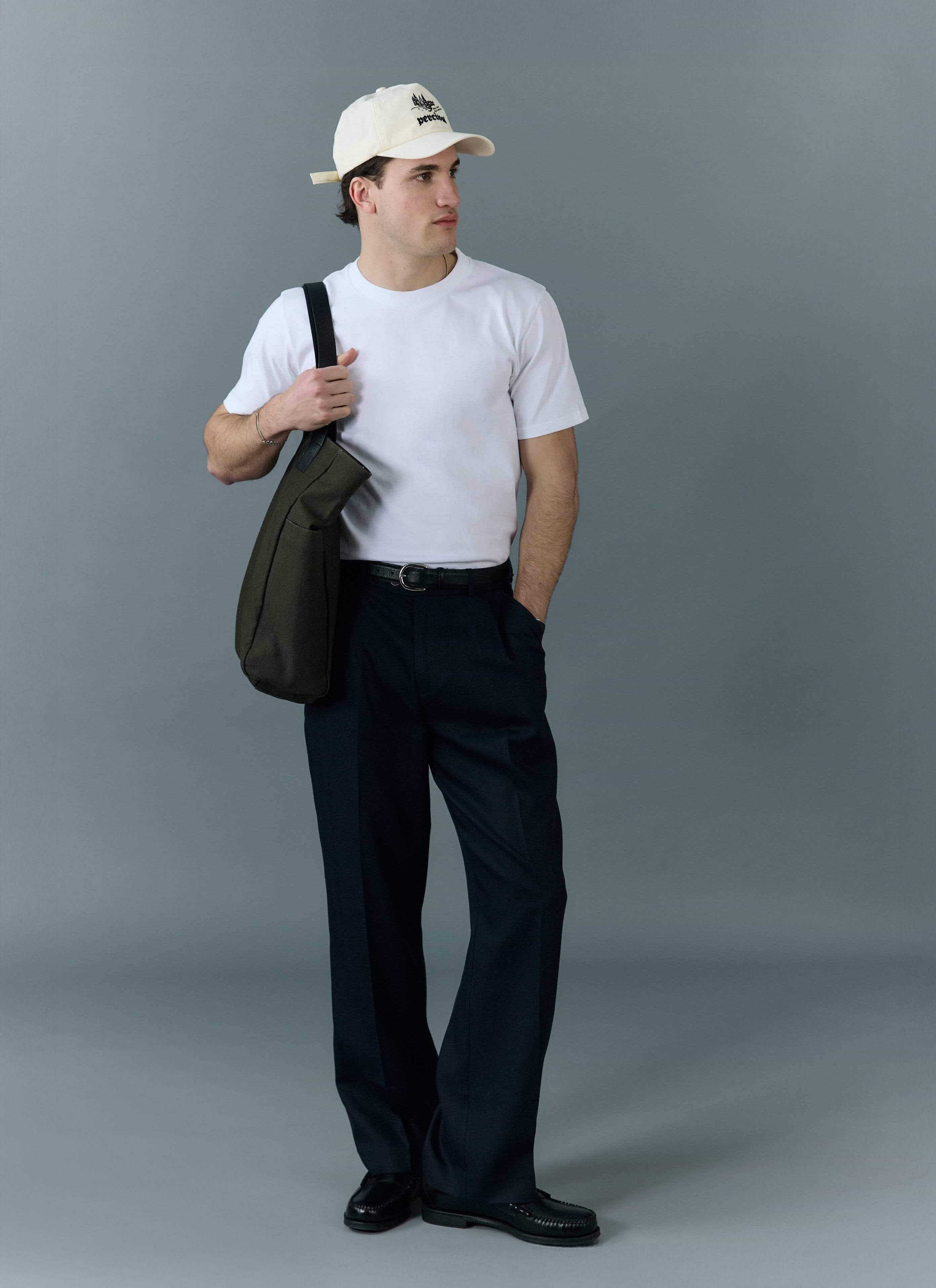 A man wearing a Percival plain white t-shirt, dark trousers, a cap, and carrying a tote bag against a grey background.
