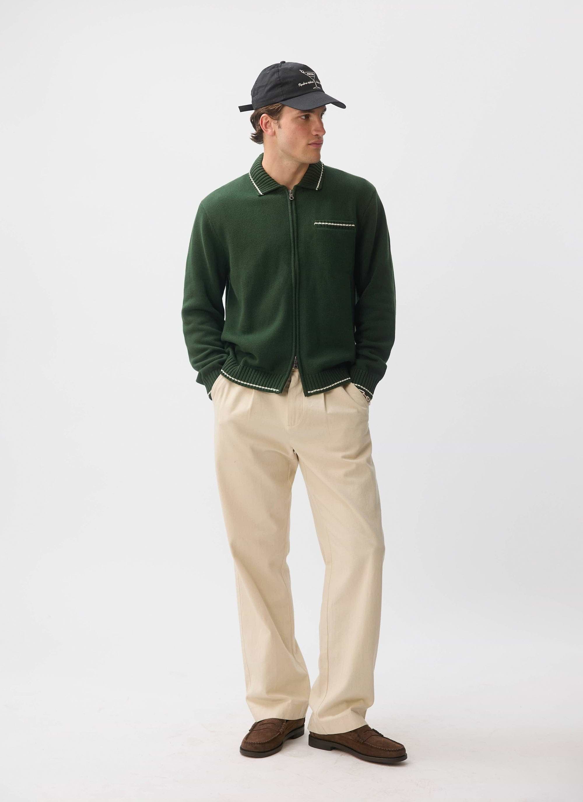 A model wears the Percival Gardner Dense Knitted Zip Cardigan in forest green, styled with cream trousers and a black cap.