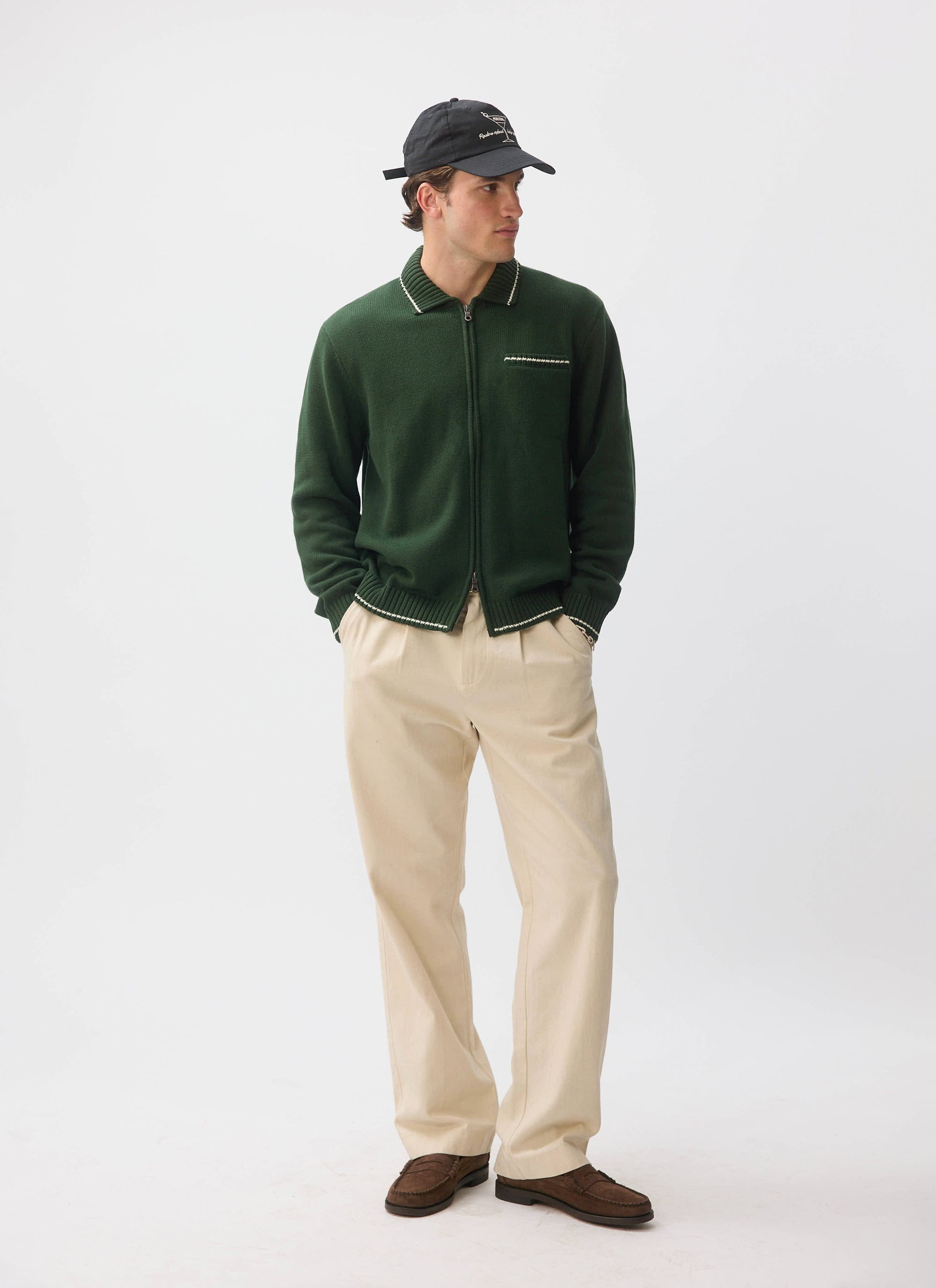 A model wears the Percival Gardner Dense Knitted Zip Cardigan in forest green, styled with cream trousers and a black cap.