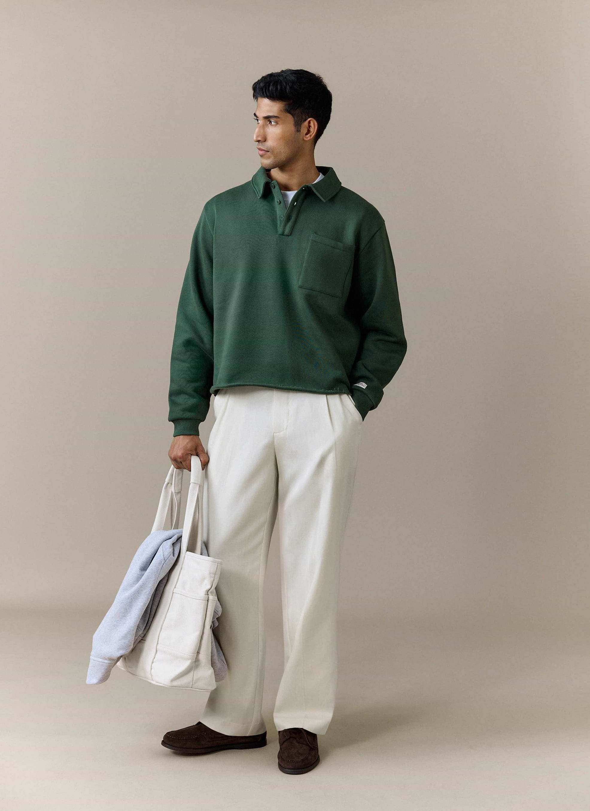 A model styles the Percival Dudley Boxy Sweatshirt in forest green with cream trousers while carrying a white tote bag.
