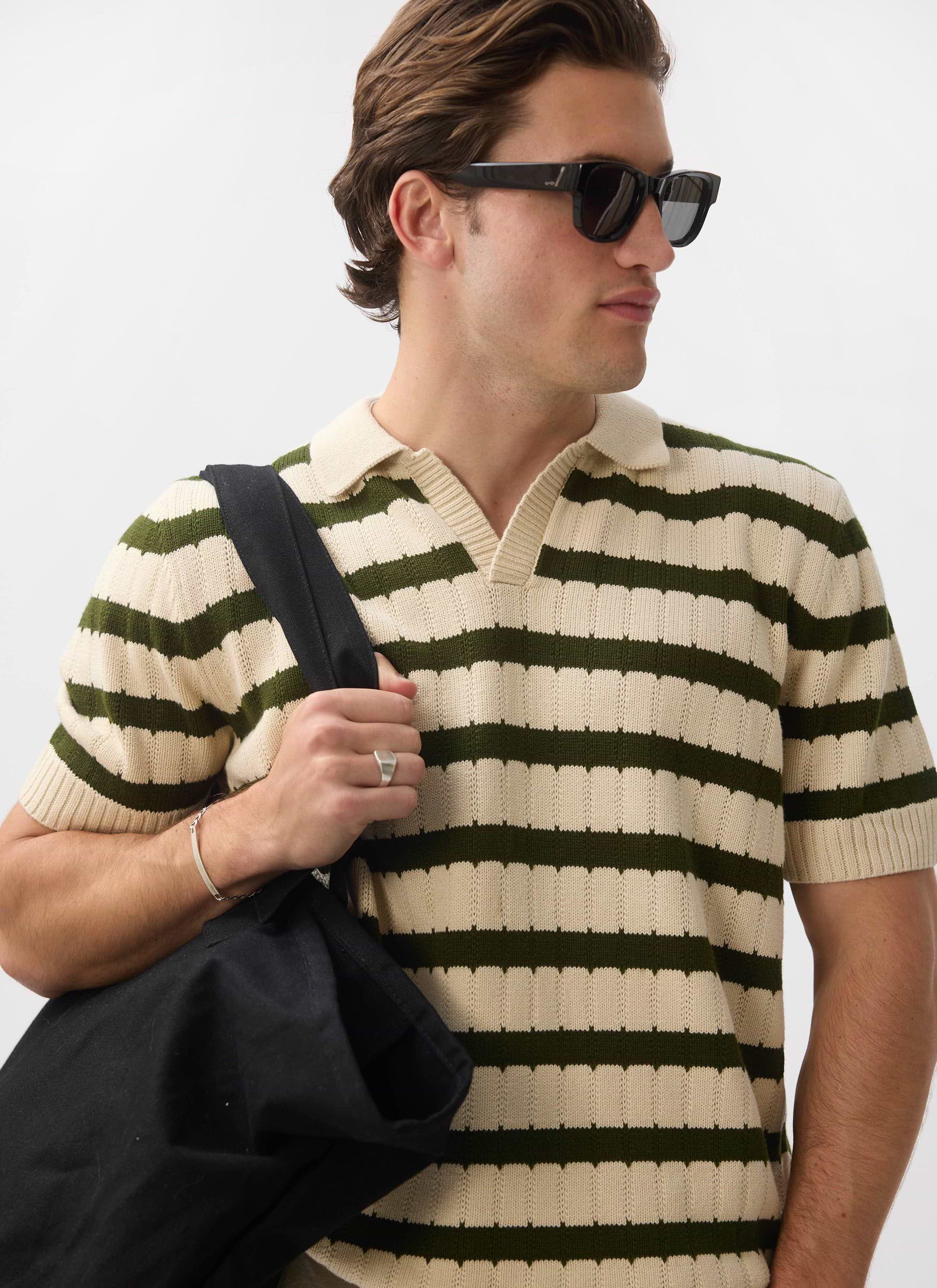 A man models the Percival Stine Knitted Polo in ecru and green, styled with sunglasses and a black tote bag.