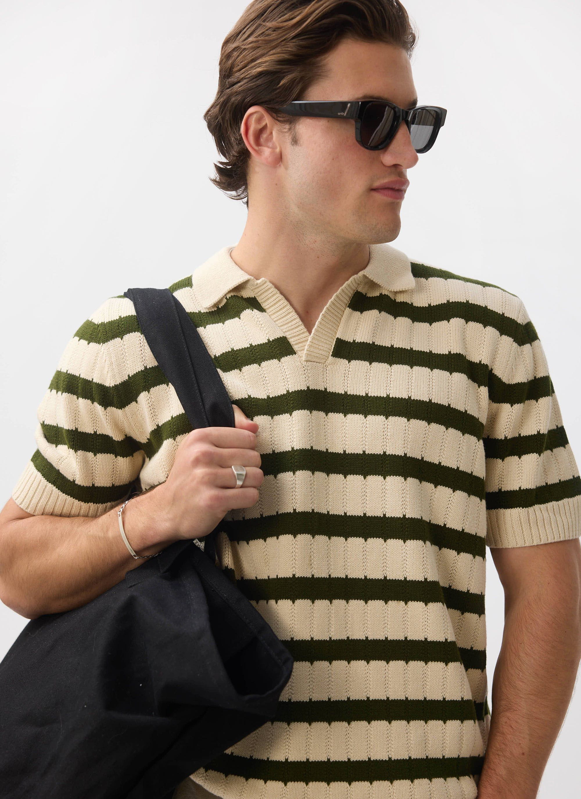 A man models the Percival Stine Knitted Polo in ecru and green, styled with sunglasses and a black tote bag.