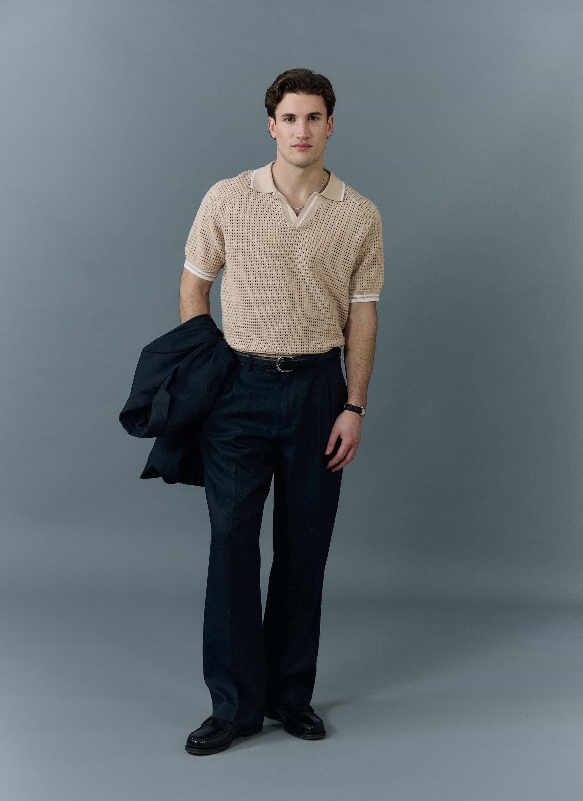 A man models the ecru Tennis Boxy Knitted Polo from Percival with dark trousers against a grey studio background.