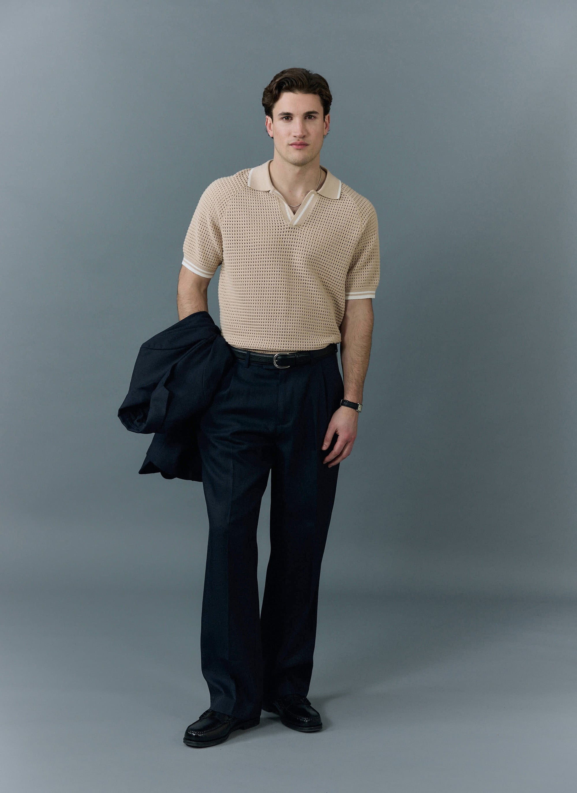 A man models the ecru Tennis Boxy Knitted Polo from Percival with dark trousers against a grey studio background.
