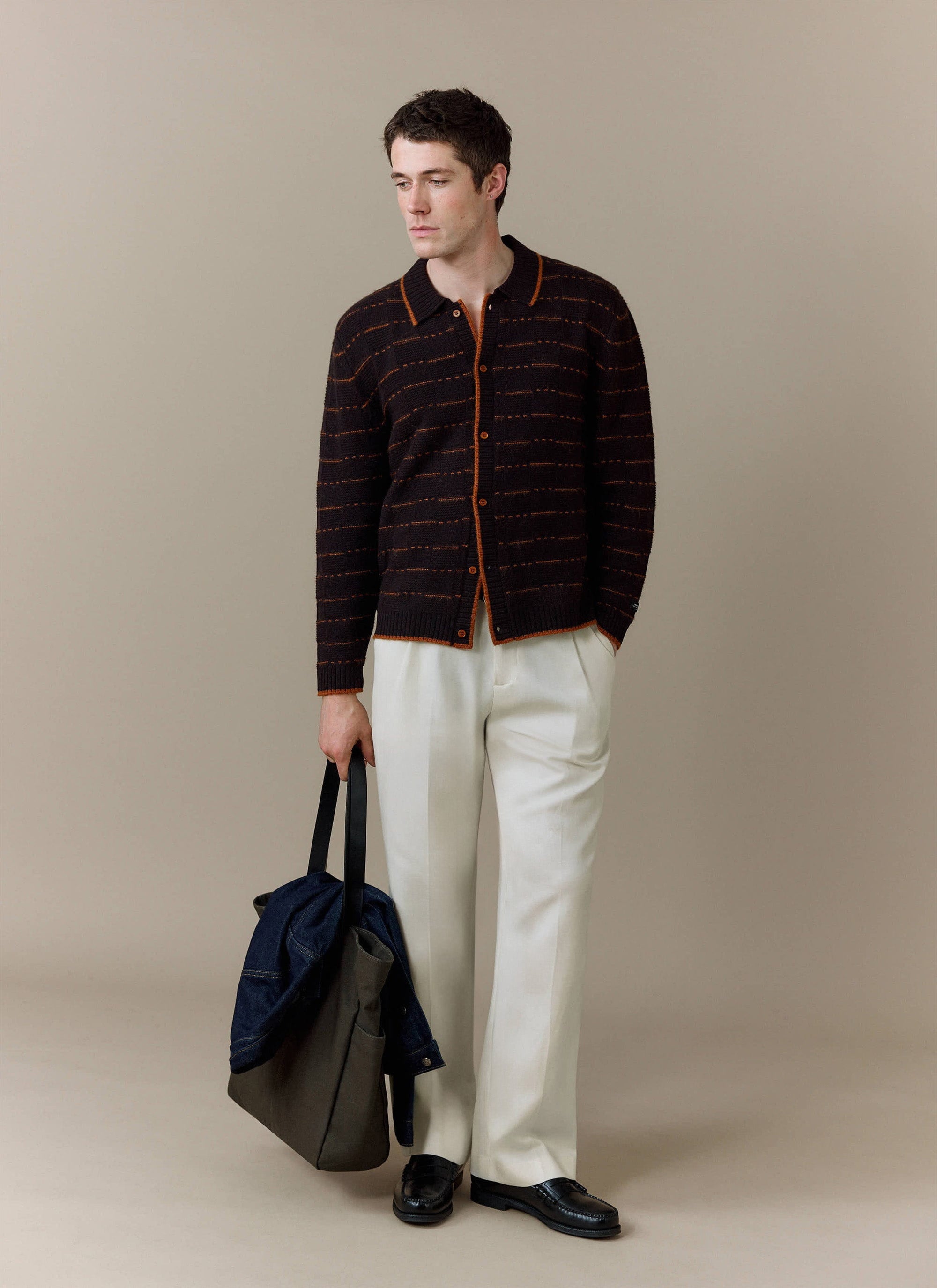 A model wears the brown Addison Knitted Shirt from Percival, styled with white trousers and carrying a tote bag.