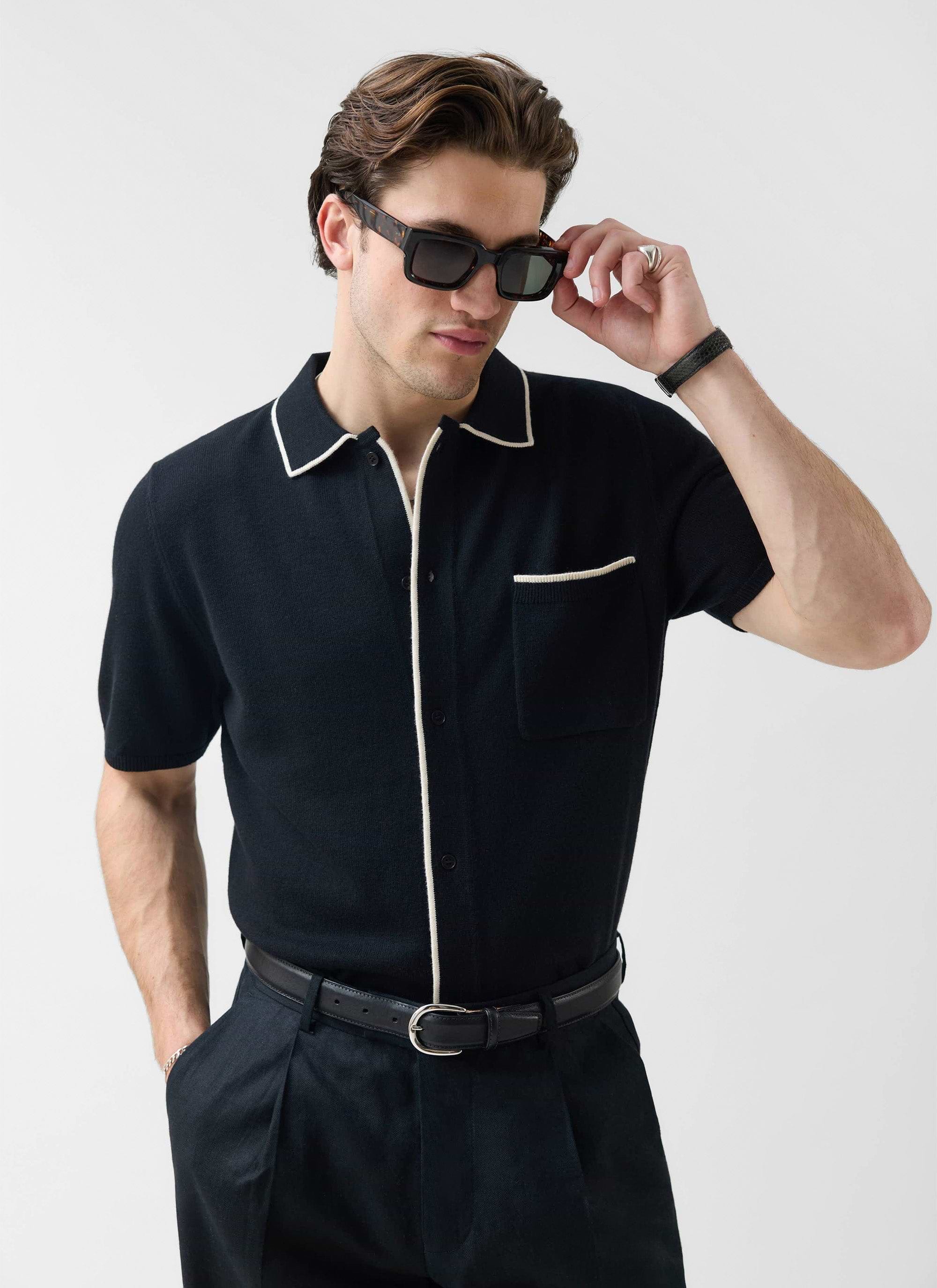 A man wearing the Percival Contrast Trim Regular Knitted Shirt in black with ecru trim, adjusting his sunglasses.
