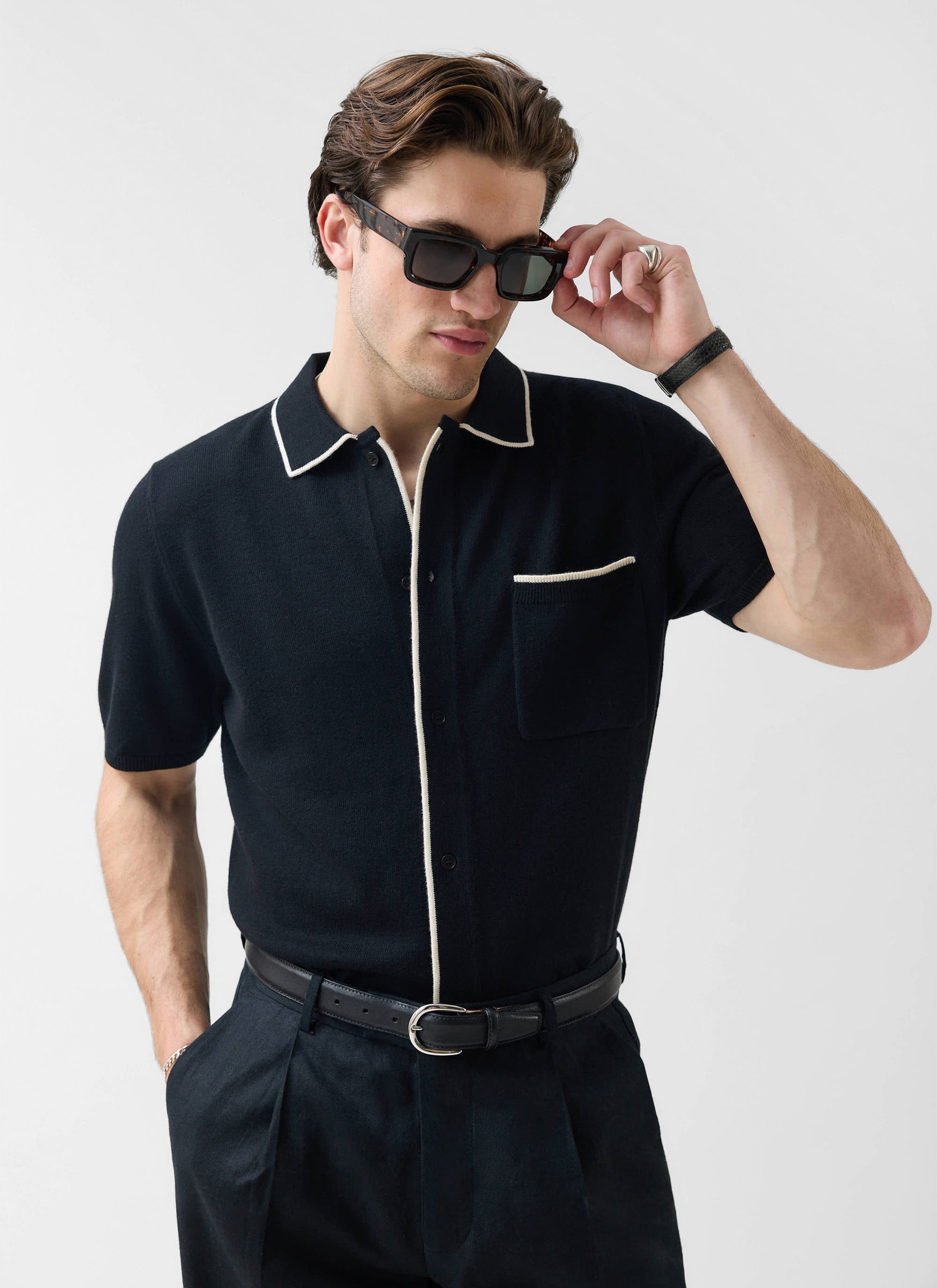 A man wearing the Percival Contrast Trim Regular Knitted Shirt in black with ecru trim, adjusting his sunglasses.