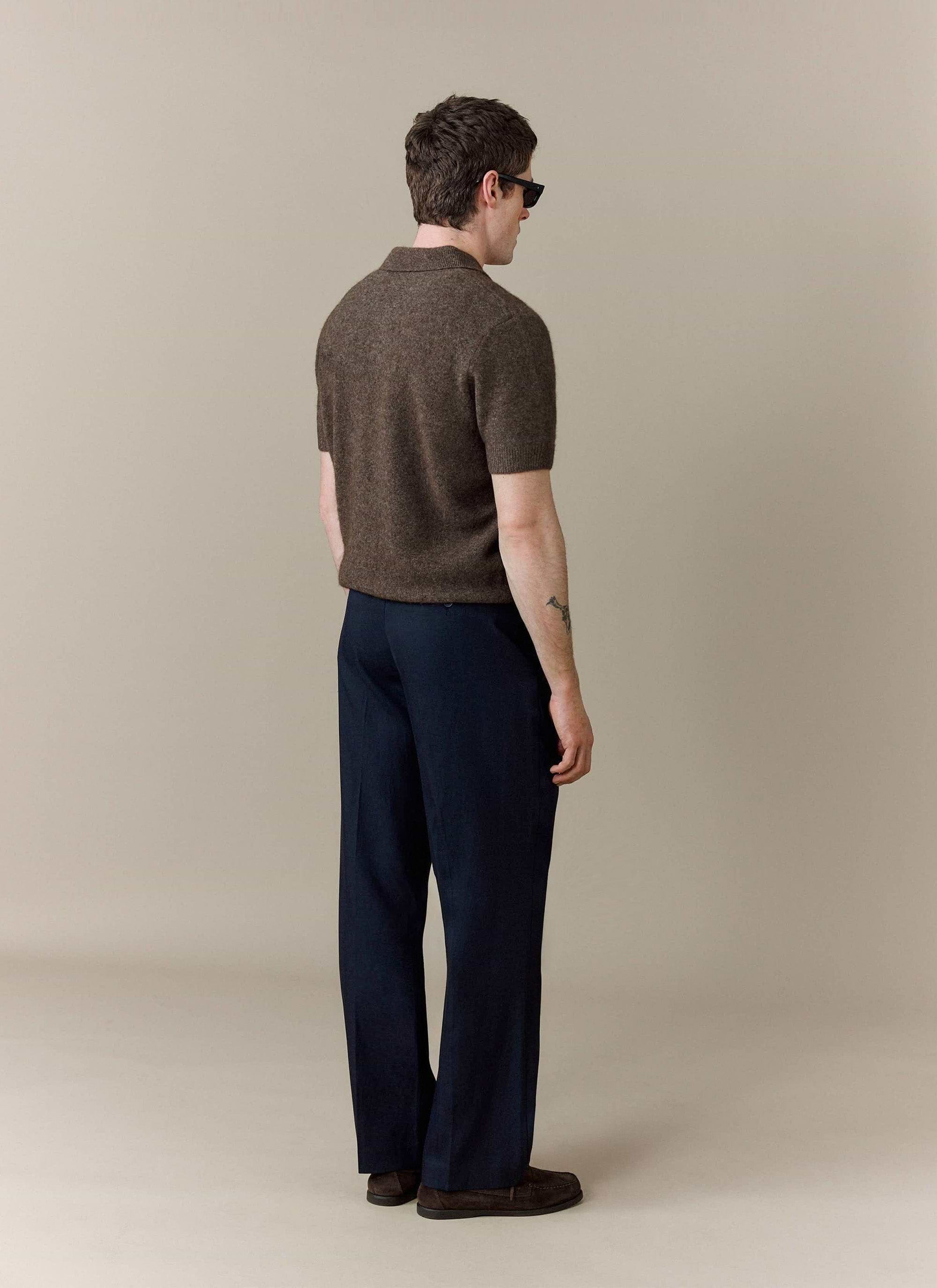 A model seen from the back wears the Percival Collins Boxy Knitted Polo in Espresso, styled with smart navy trousers.