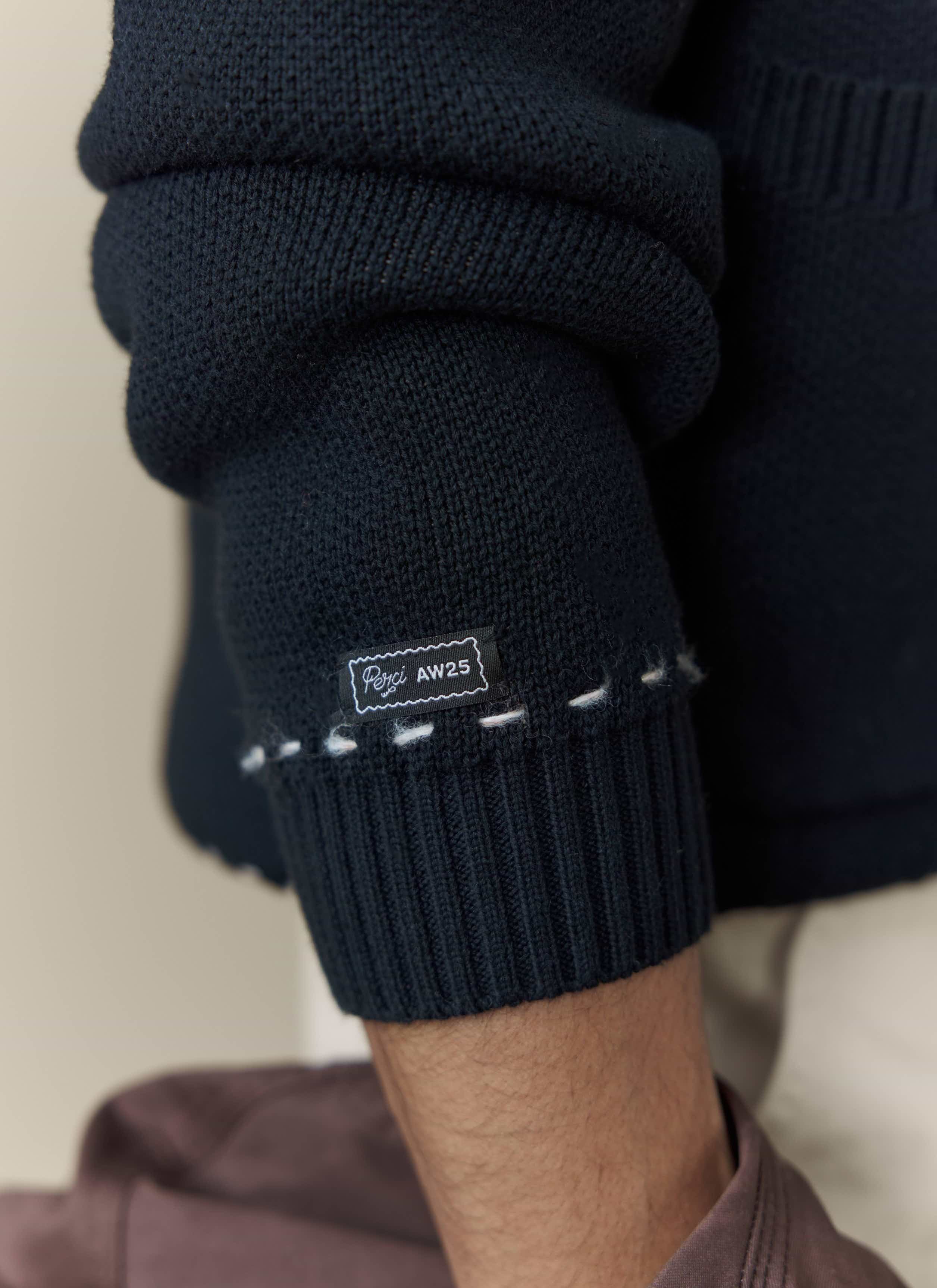 A close-up shot of the cuff on the black Percival Module Boxy Cardigan, showing the brand label and decorative stitching.