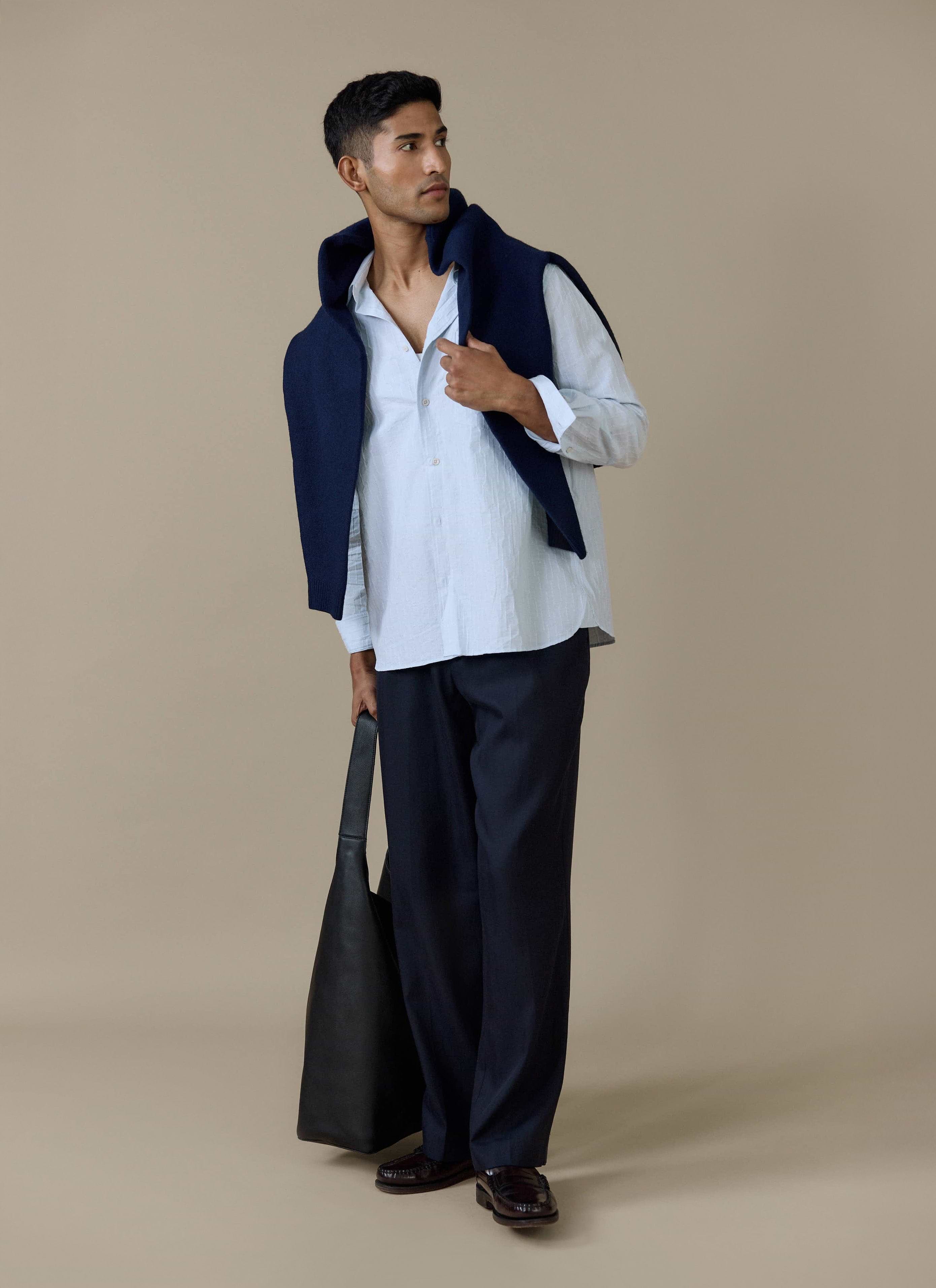 Model wears the light blue Percival Paros Pearce Shirt in seersucker, styled with navy trousers and a navy sweater.